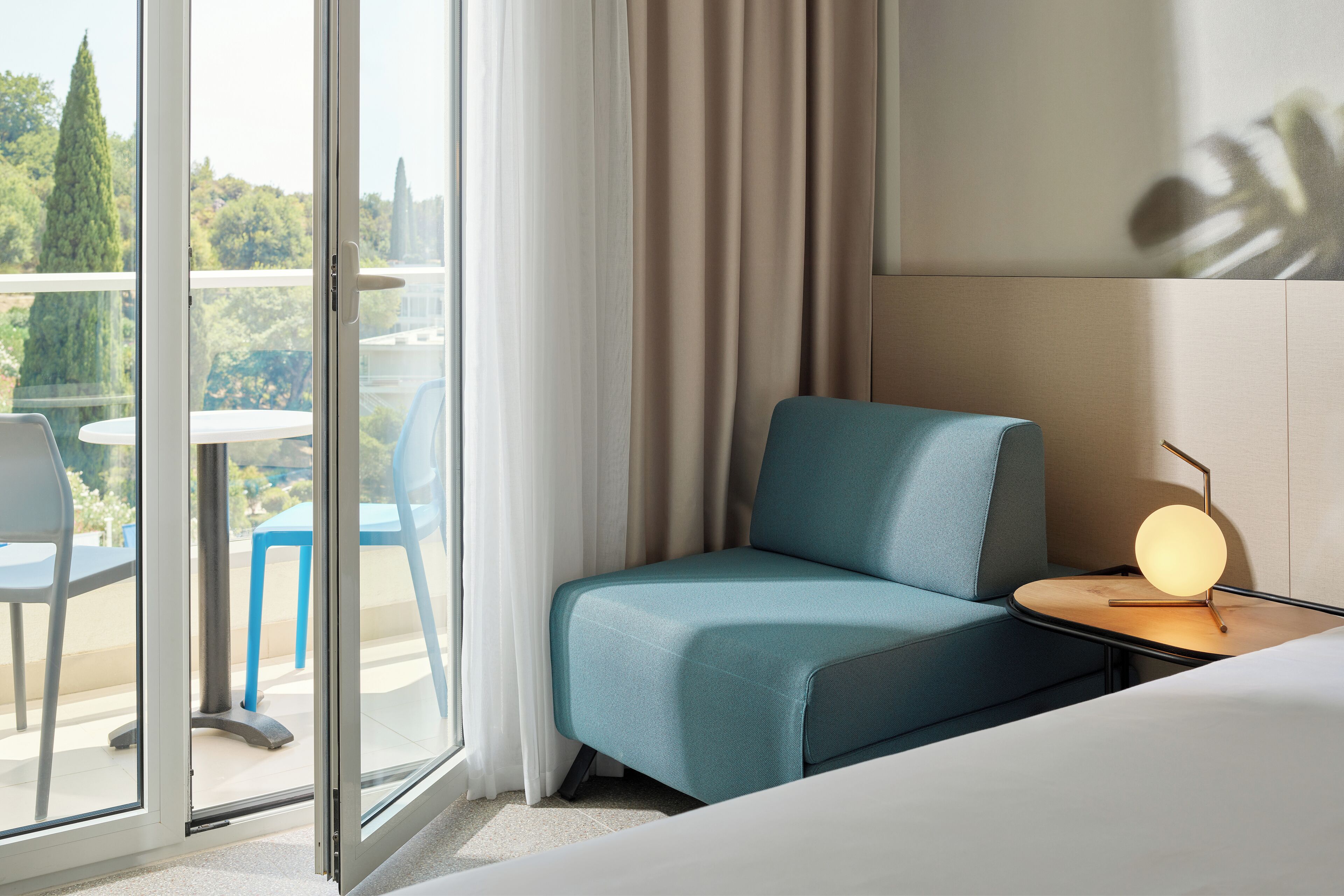 Hotelkamer met een blauwe bank, een tafeltje met een lamp en uitzicht op het landschap door een glazen deur.