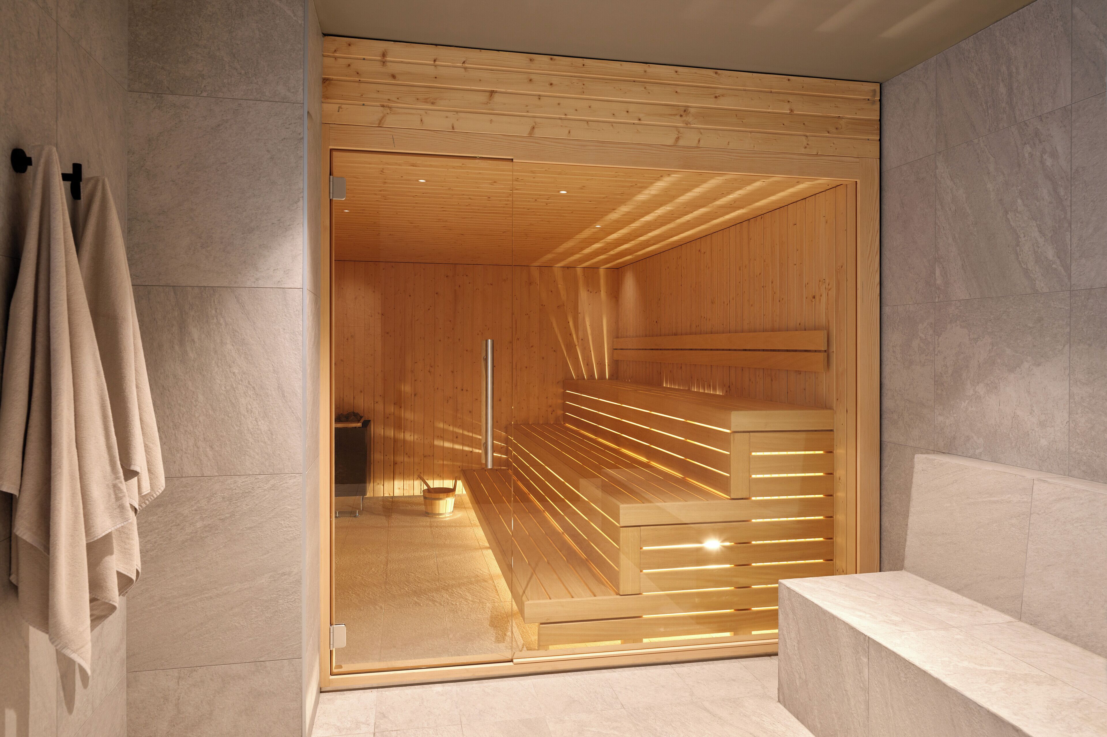 Una sauna con bancos de madera y una puerta de vidrio, frente a una pared revestida con azulejos.