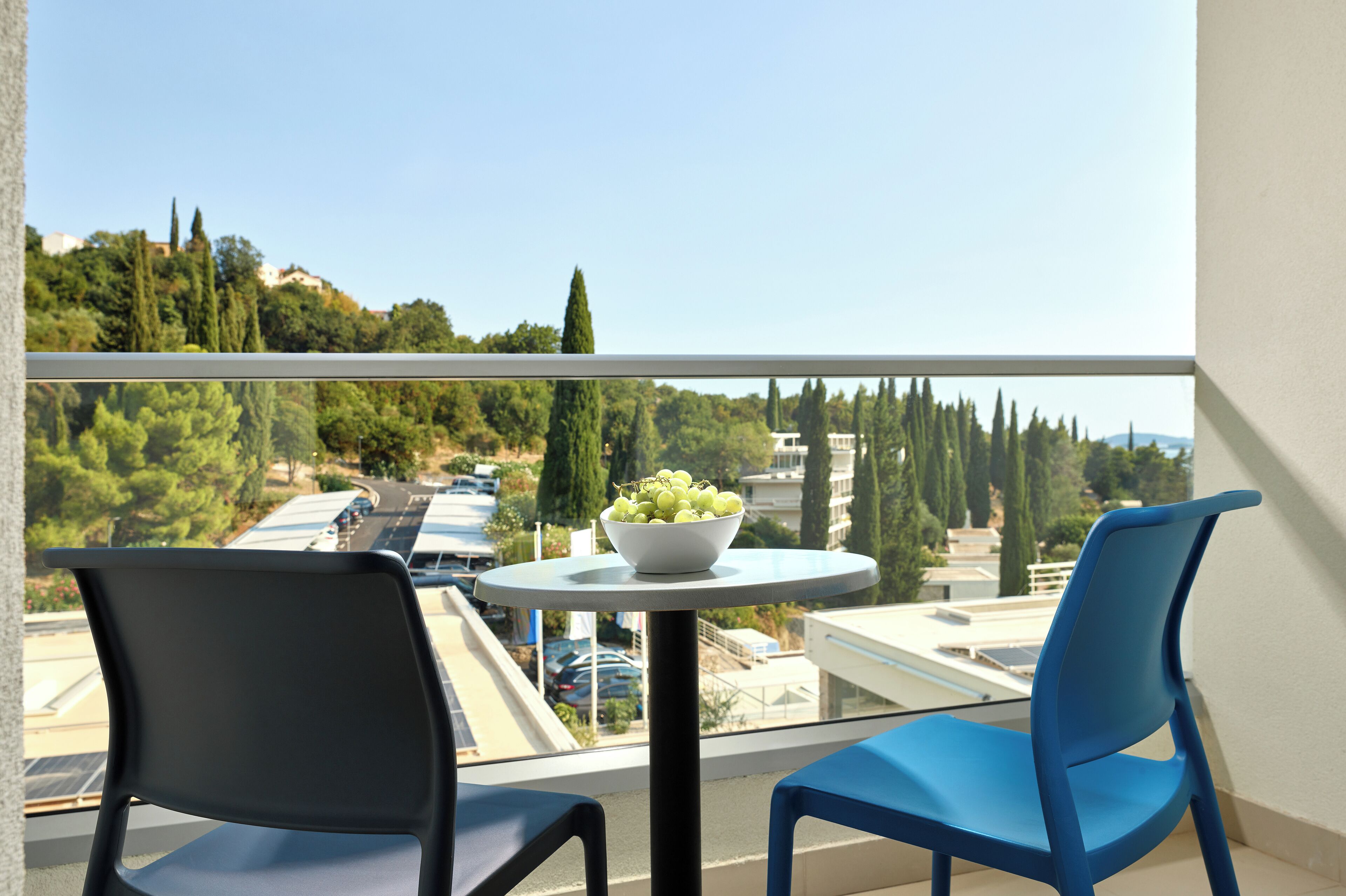 A balcony with two chairs, a table with a bowl of fruit, and a view of a parking area and trees.