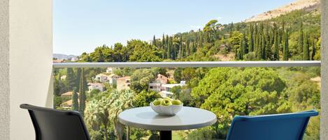 Uma varanda com duas cadeiras, uma mesa com uma tigela de frutas e uma vista panorâmica de uma paisagem verde com árvores e edifícios.
