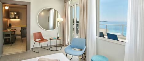 A hotel room with a bed, a round mirror, two chairs, a small table, and a view of the sea through large windows.