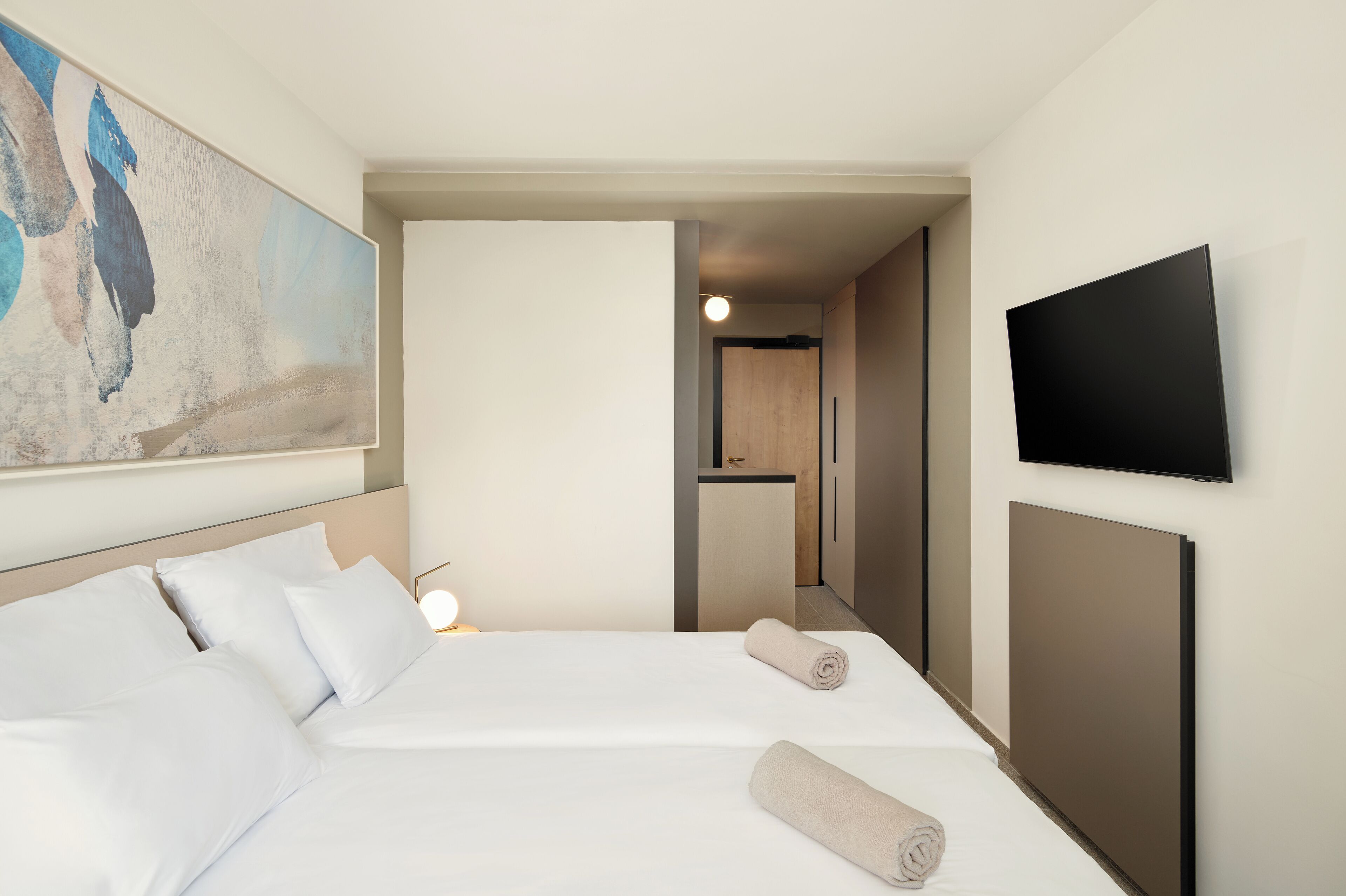 A hotel room with a bed, a TV mounted on the wall, and a painting on the wall.