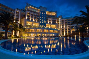Un hotel grande y moderno con piscina central, múltiples balcones y una fachada bien iluminada.