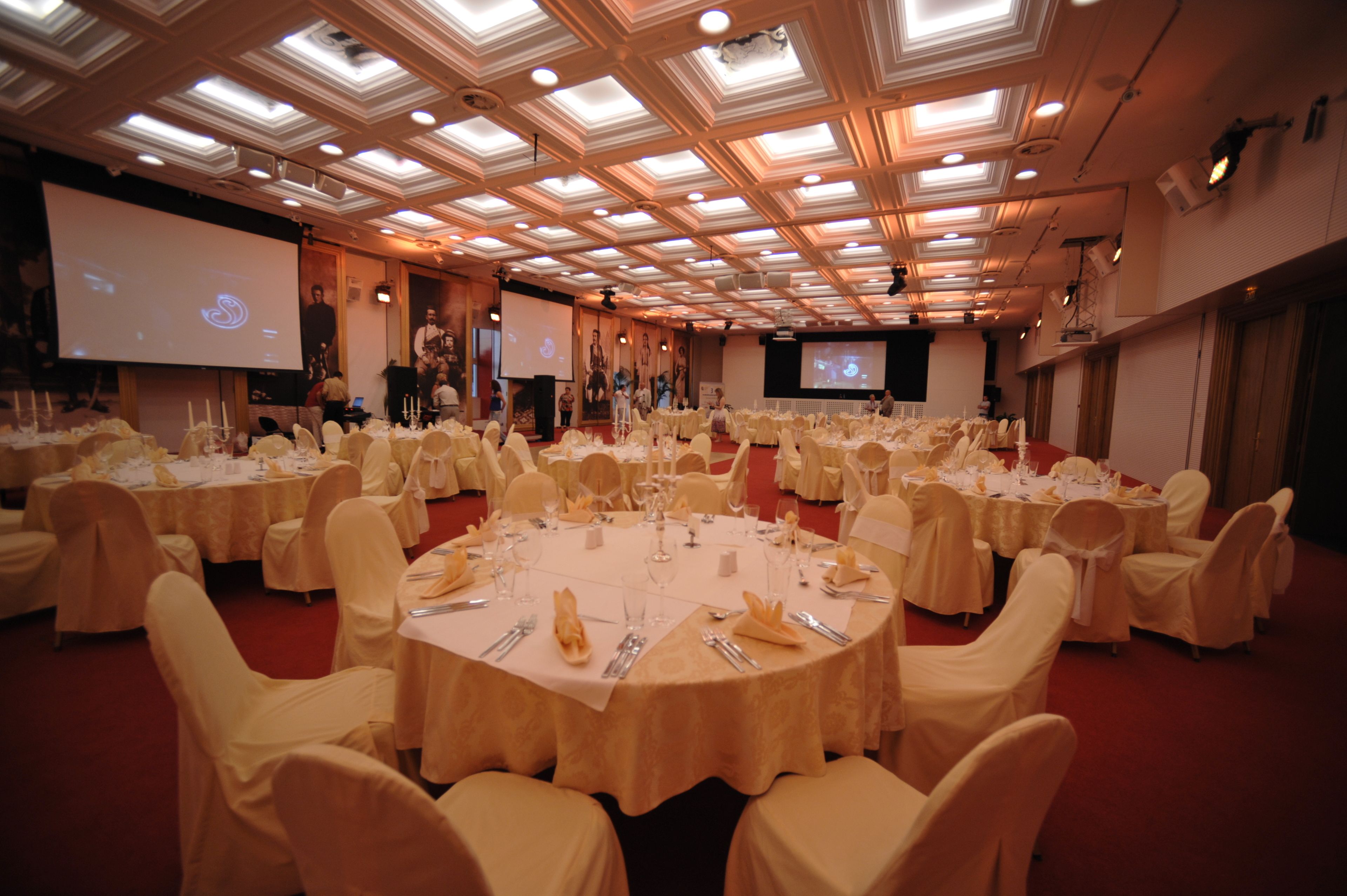 A large banquet hall with round tables set for a formal event, featuring a high ceiling with recessed lighting and a projection screen.