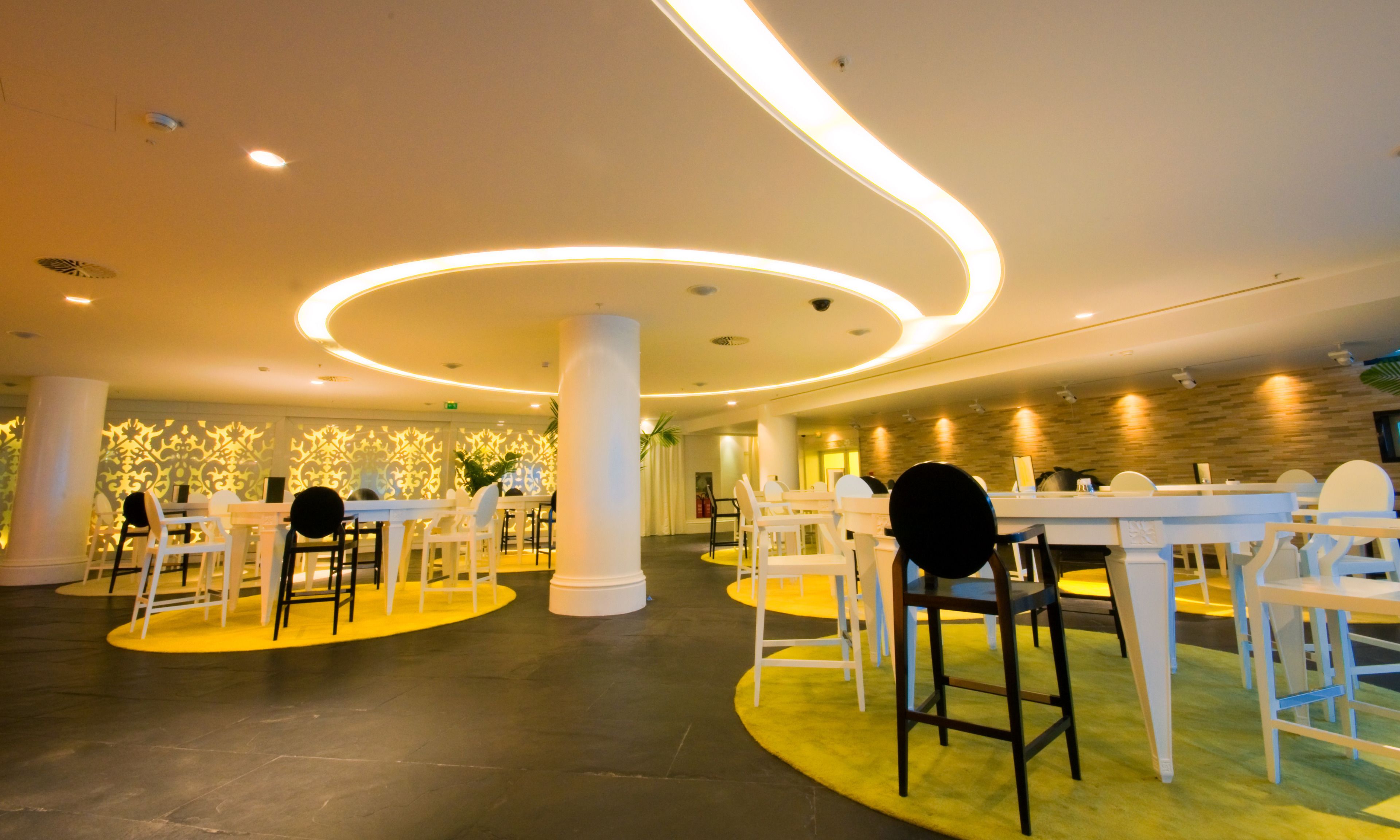 A spacious indoor dining area with white tables and chairs, a modern ceiling design with circular lighting, and a patterned wall in the background.