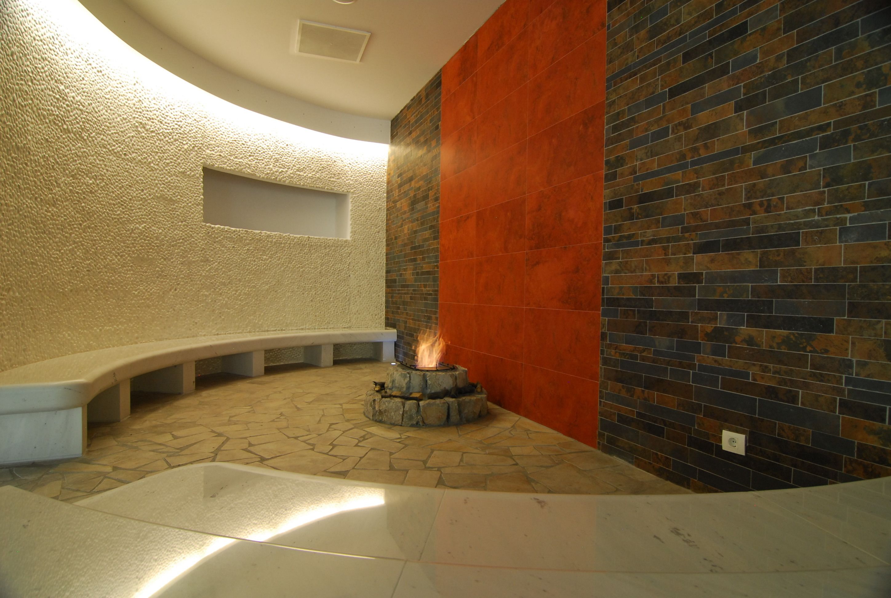 Un espacio circular con asientos, una chimenea, suelo de piedra y baldosas de pared contrastantes.