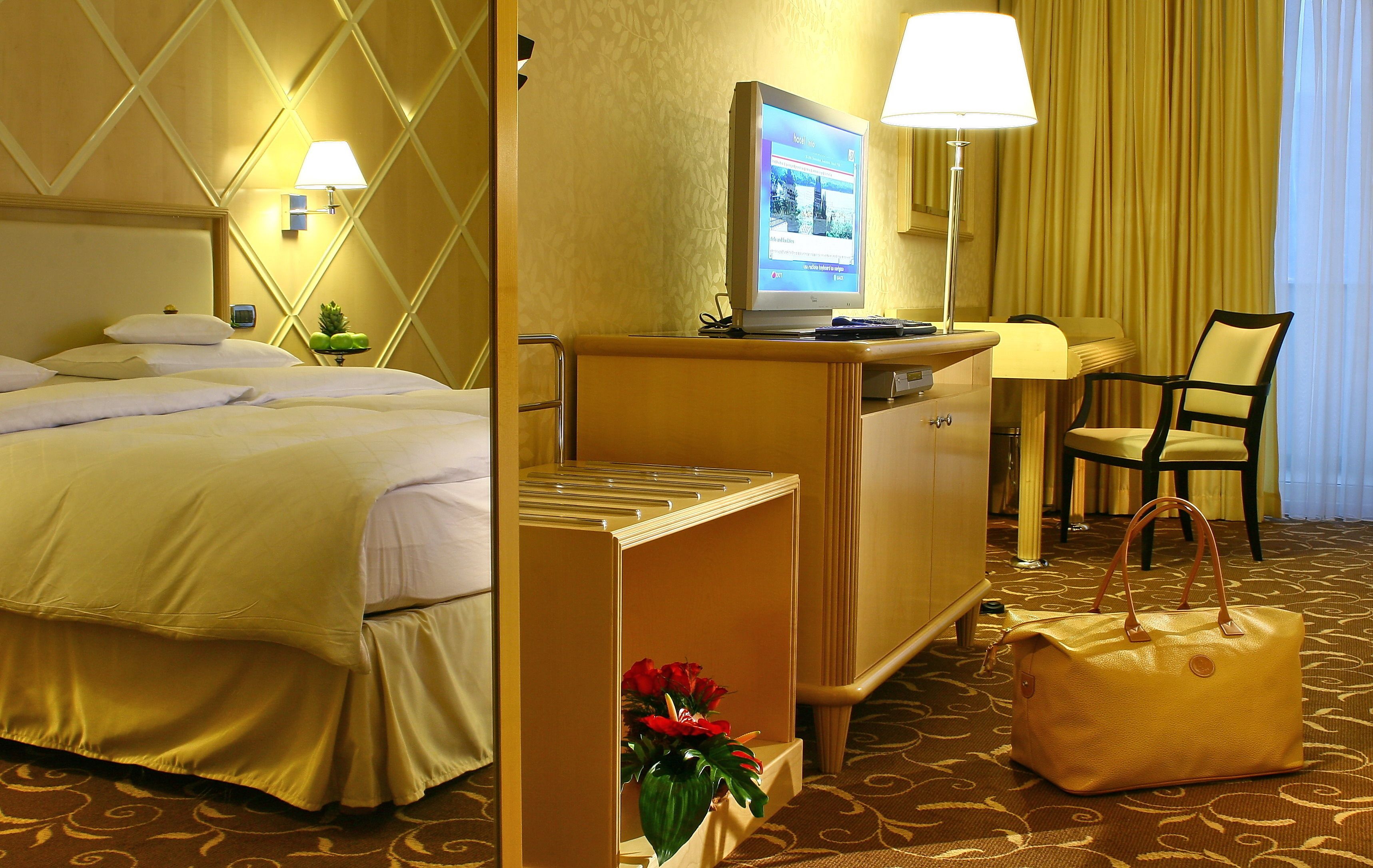 A hotel room with a bed, a desk with a computer, a chair, and a handbag.