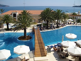 Zona de piscina con sillas de descanso y sombrillas, rodeada de palmeras y vista al mar.