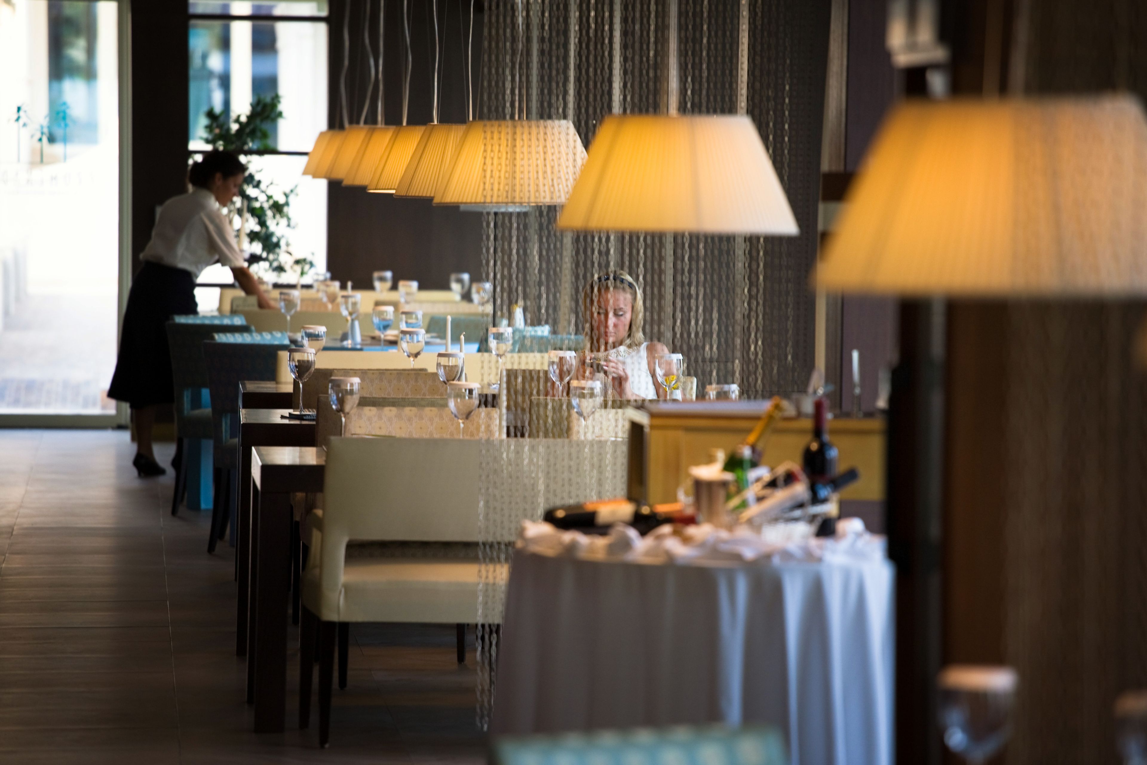 Un restaurante con mesas dispuestas para comer, un camarero al fondo y lámparas colgantes.