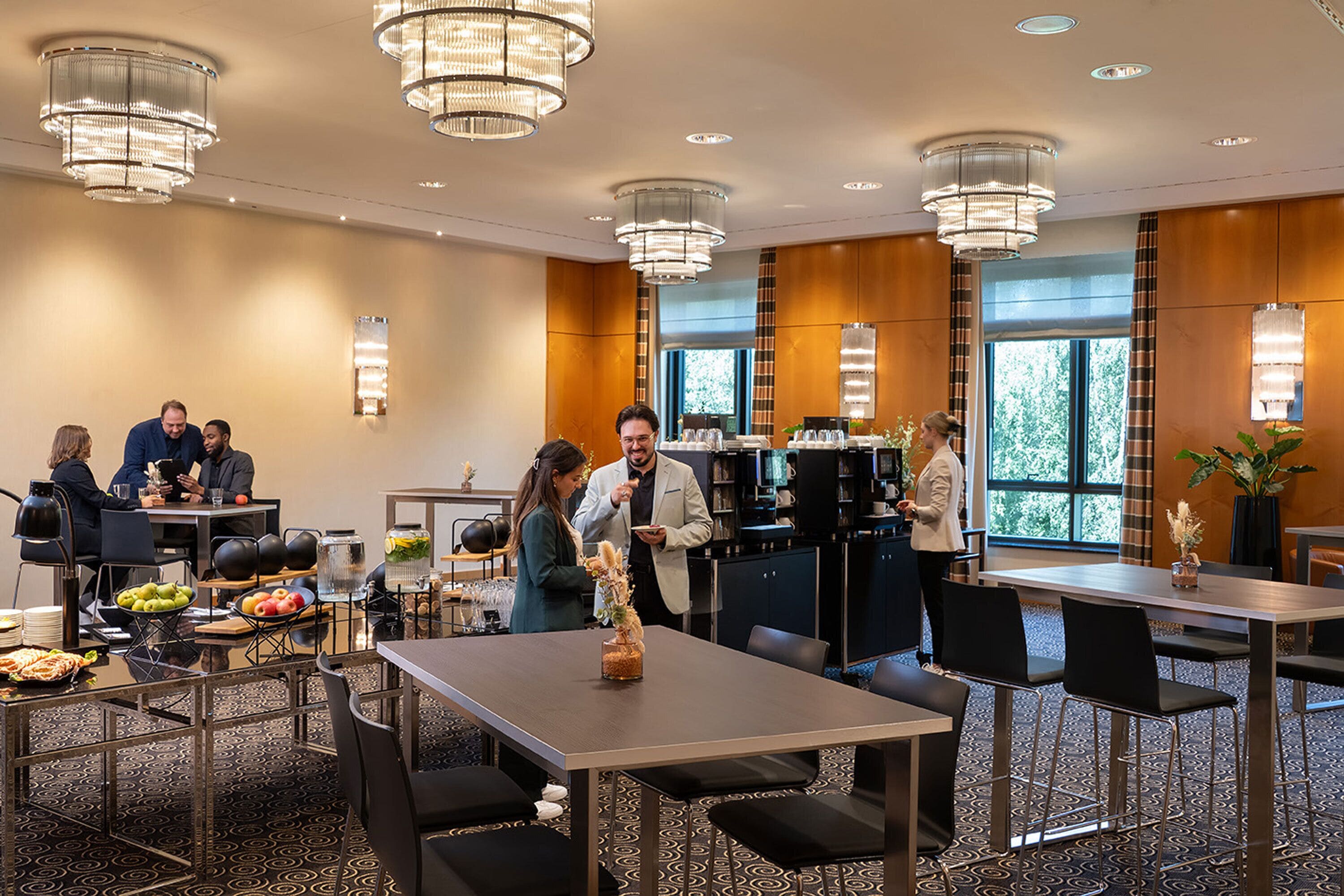 Een restaurantgedeelte van een hotel met meerdere tafels, stoelen en kroonluchters. Gasten dineren en maken er gezellig praatje.