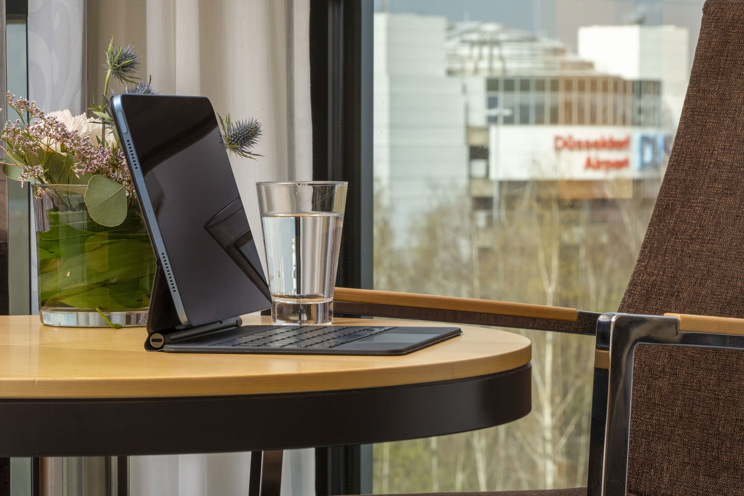 Um laptop sobre uma mesa, com um copo de água, um vaso de flores e vista para um aeroporto ao fundo.