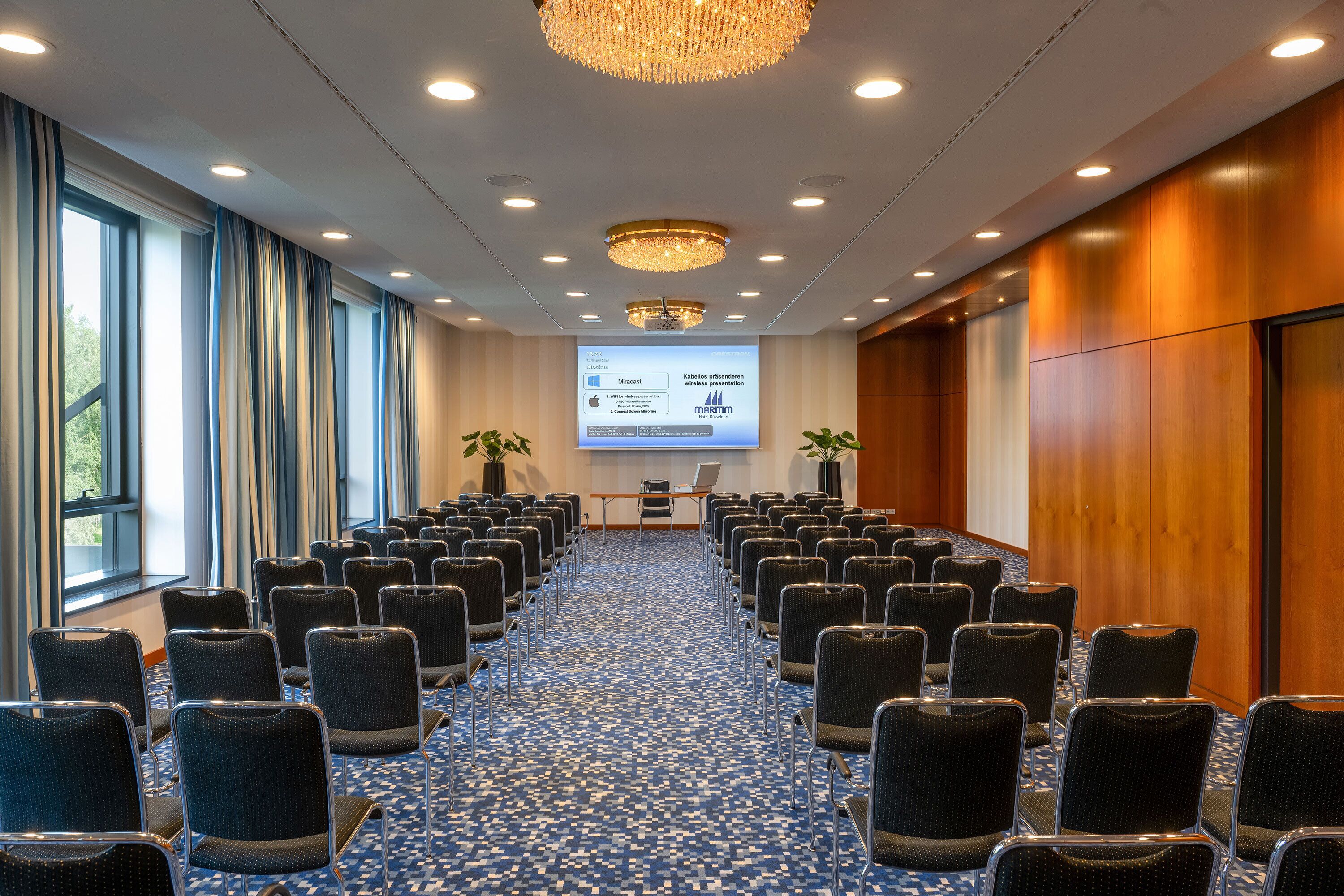 Un salón de conferencias con filas de sillas, una pantalla de proyector y un amplio ventanal con cortinas.