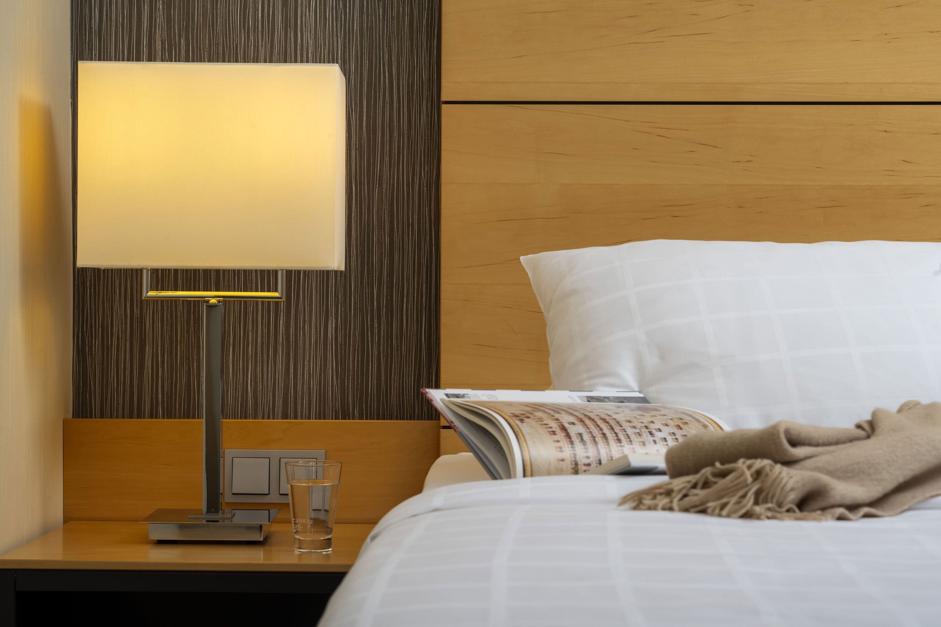 Una cama bien hecha con un almohadón blanco y una manta color beige, una mesita de noche con una lámpara, un vaso de agua y una revista.