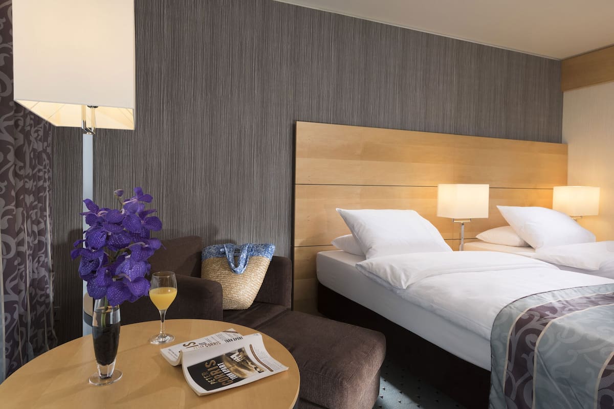 A hotel room with a bed, bedside lamps, a table with a newspaper, a vase with purple flowers, and a chair.