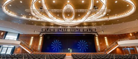 Un amplio auditorio con hileras de sillas, un escenario con fondo azul y una imponente lámpara de cristal.