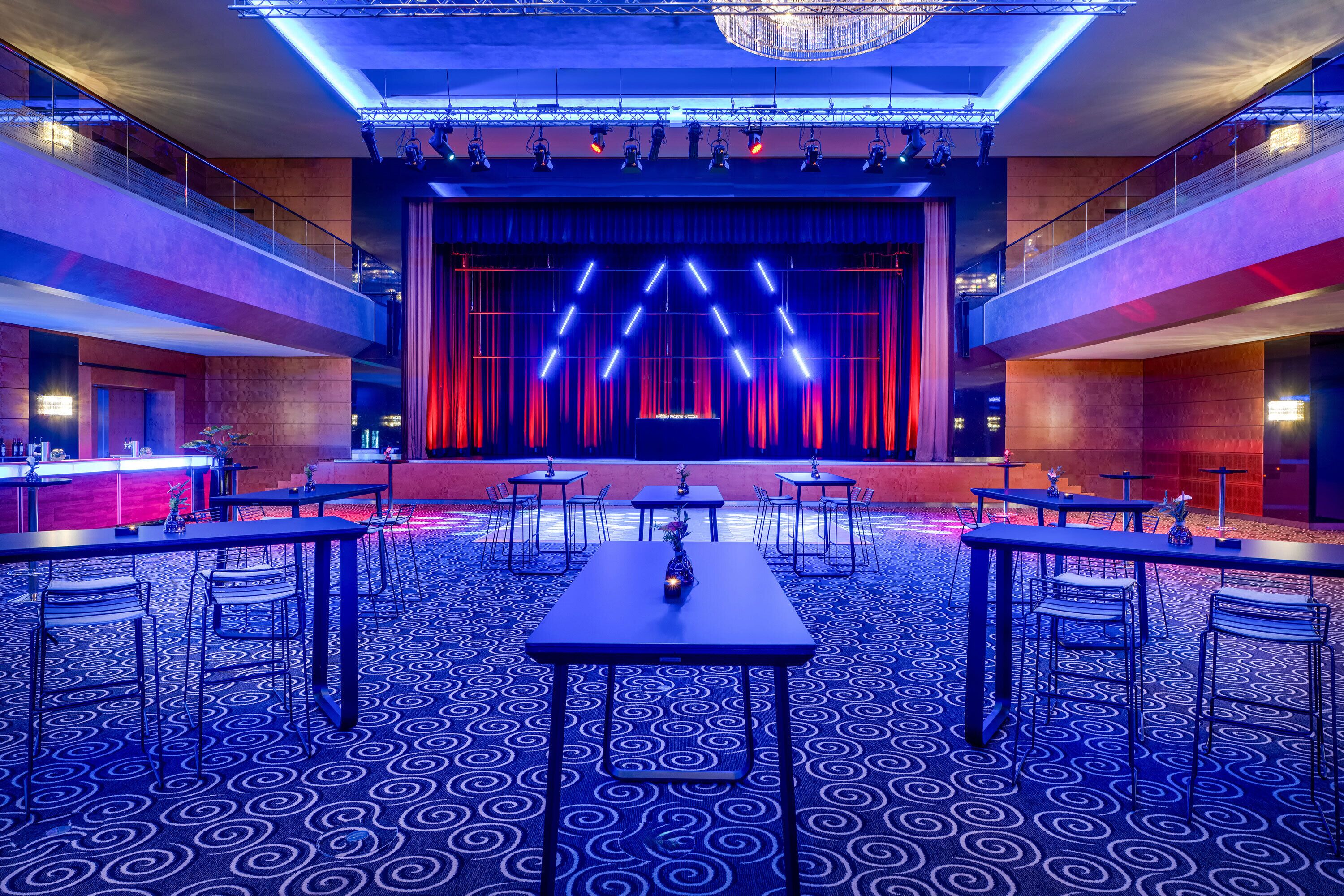 Un salón de conferencias moderno con un escenario, mesas y sillas dispuestas para presentaciones.