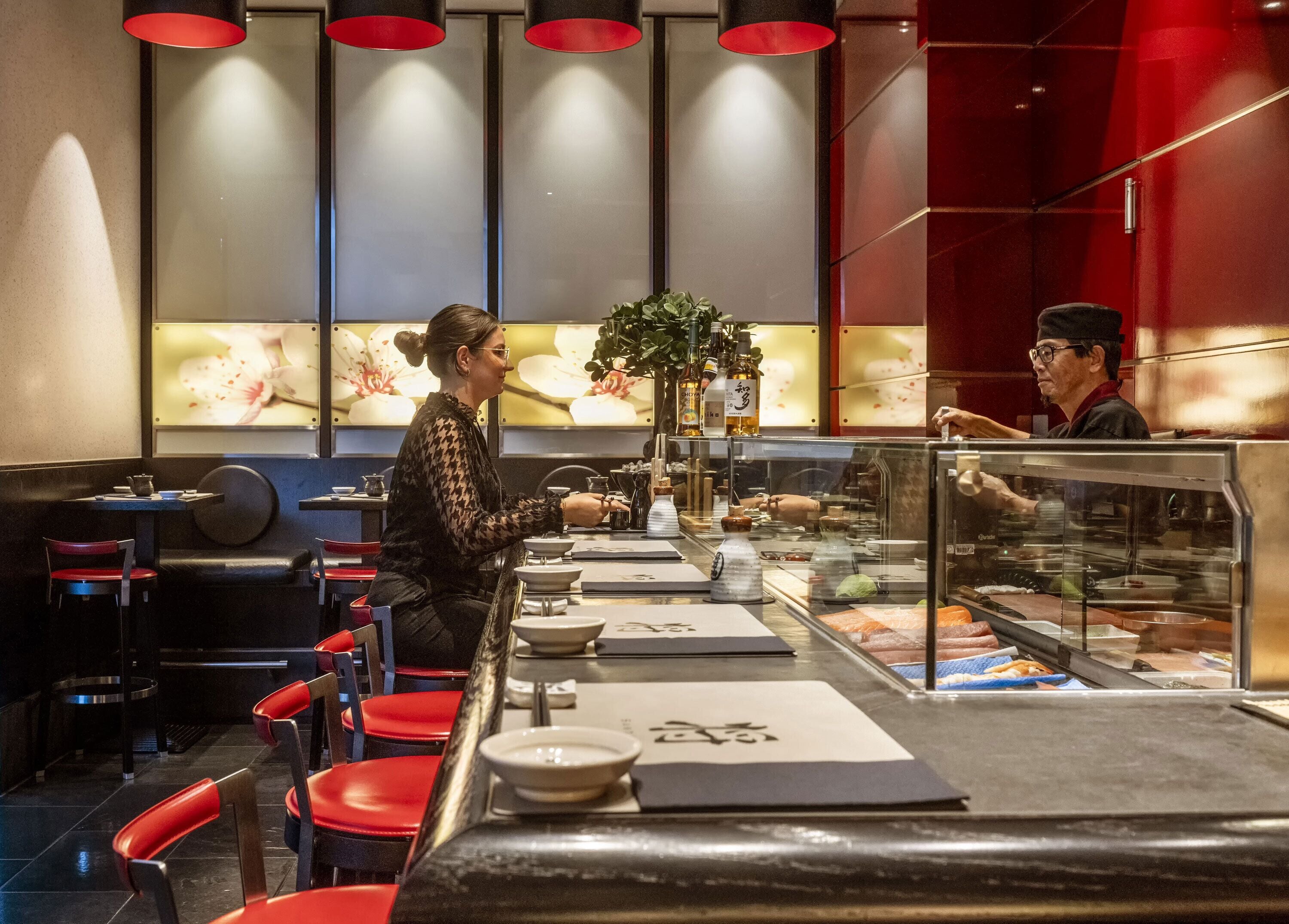 L’intérieur d’un restaurant avec un bar à sushis, des chaises rouges et un comptoir où un serveur s’occupe d’un client.