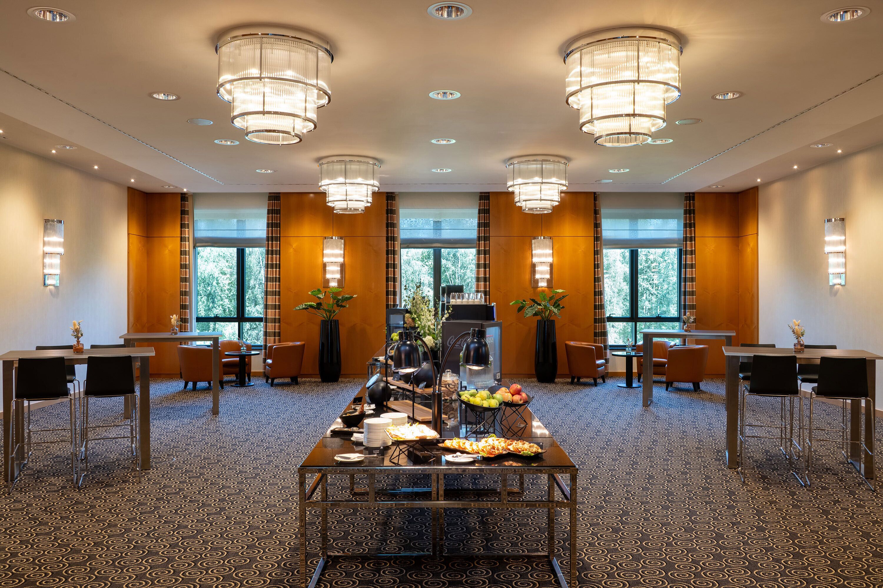 Un salón de conferencias de hotel con una larga mesa dispuesta para un bufé, múltiples sillas y grandes ventanas con cortinas.