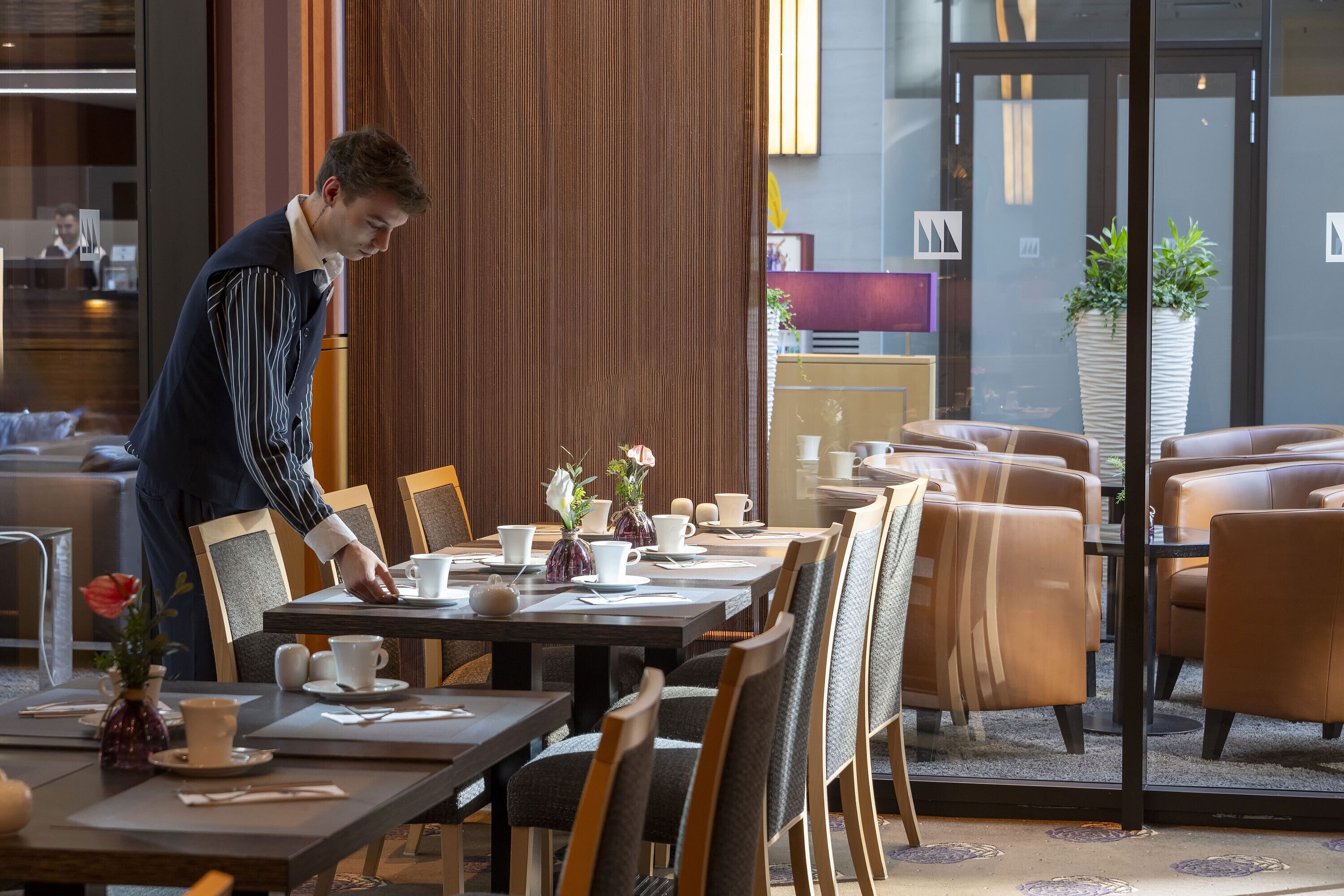 Un comedor de hotel con mesas dispuestas para el servicio, una recepción y una planta grande en maceta.