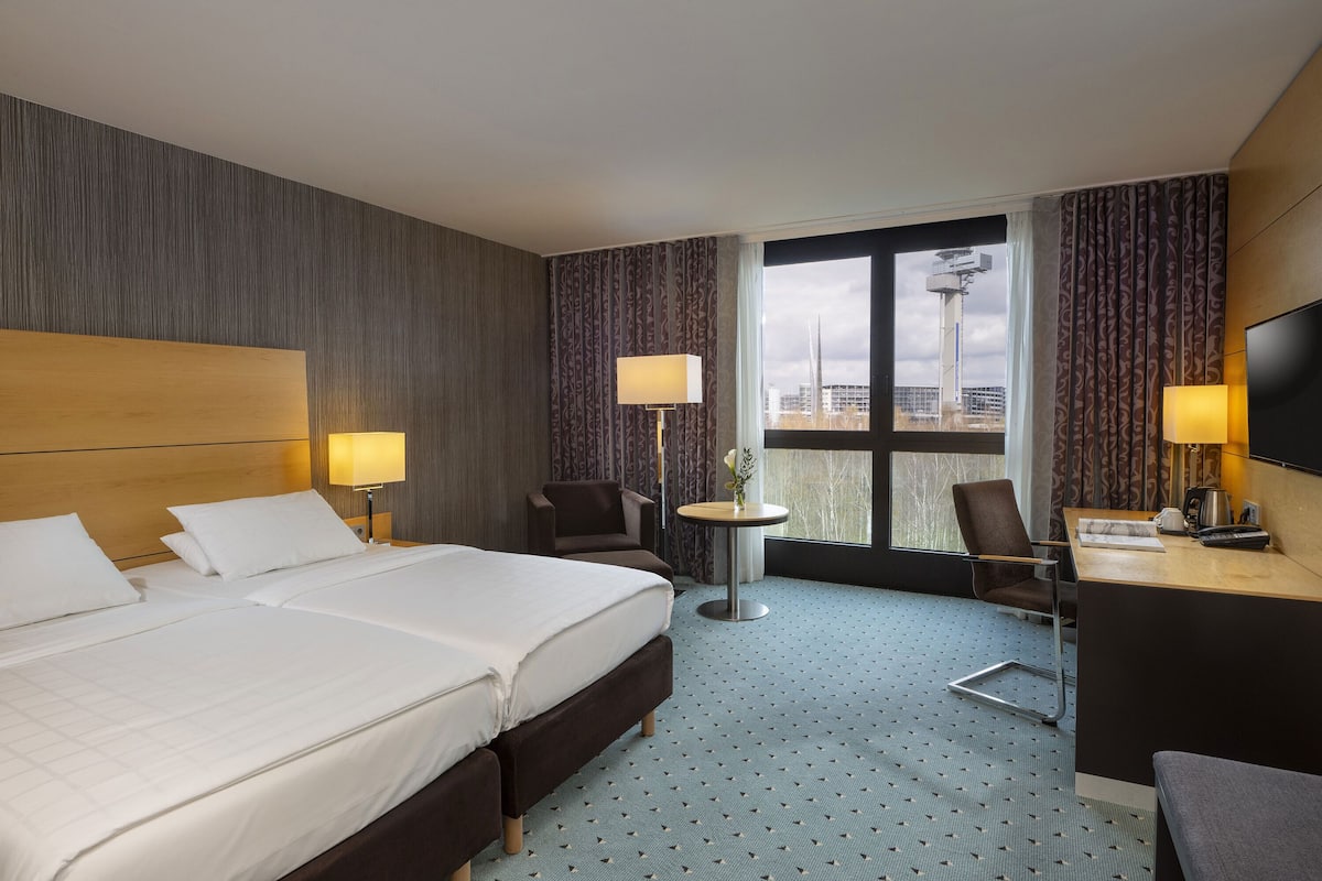 A hotel room with two beds, a desk, a chair, a TV, and a window with a view of a building.