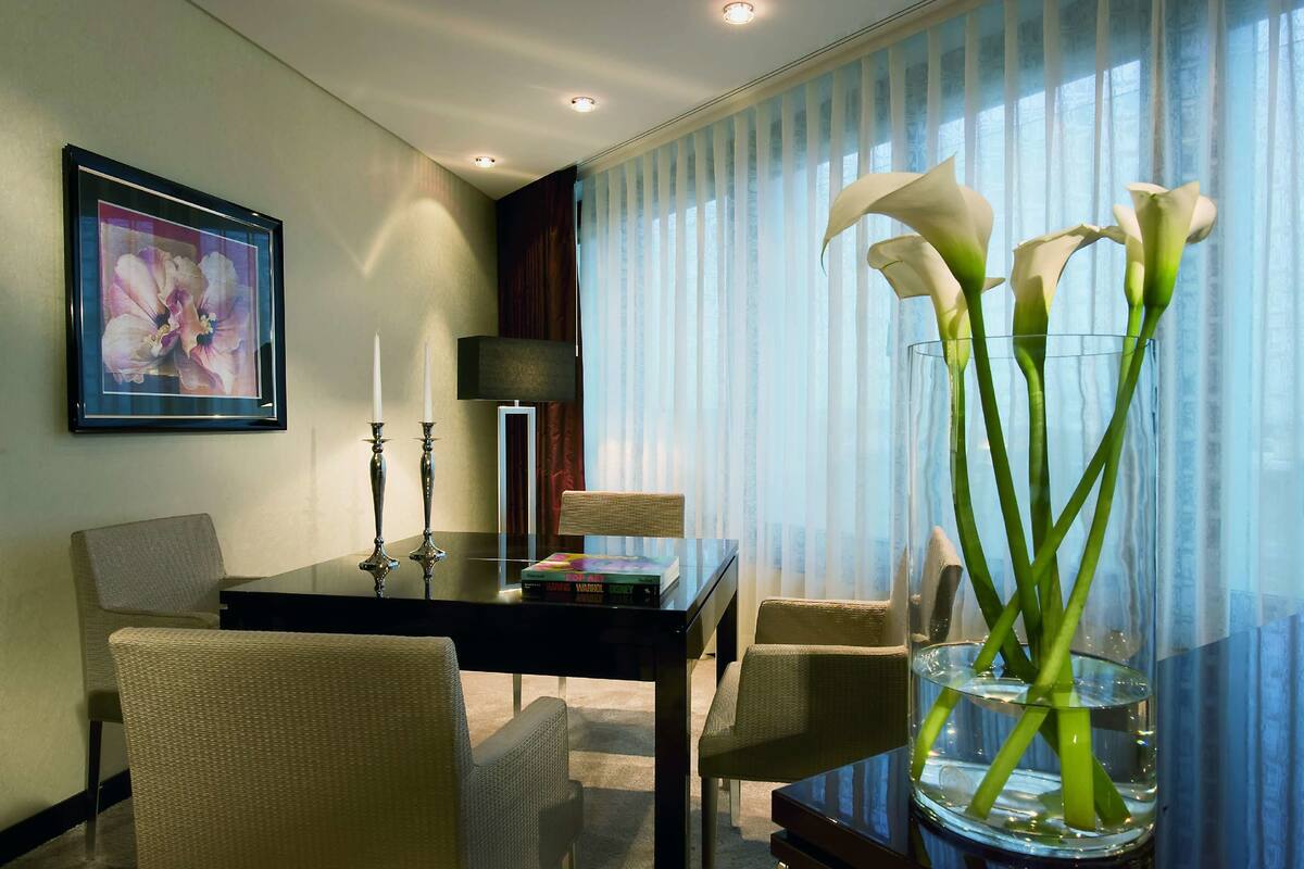 A hotel room with a dining table, chairs, a framed floral artwork, a lamp, and a vase with calla lilies.