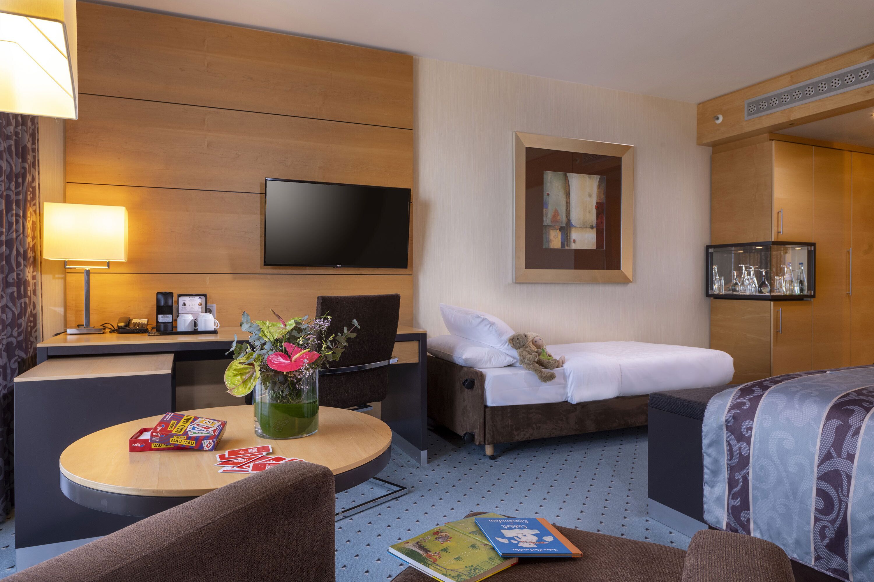 A hotel room with a bed, a desk with a lamp, a TV, a chair, a small table with a vase of flowers, and a bookshelf.