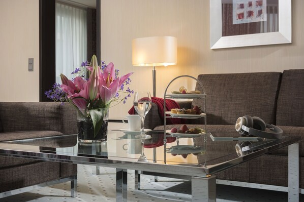 A modern living room with a glass coffee table, a vase of flowers, a lamp, a sofa, and a tray with pastries. - Maritim Hotel Düsseldorf (Düsseldorf)
