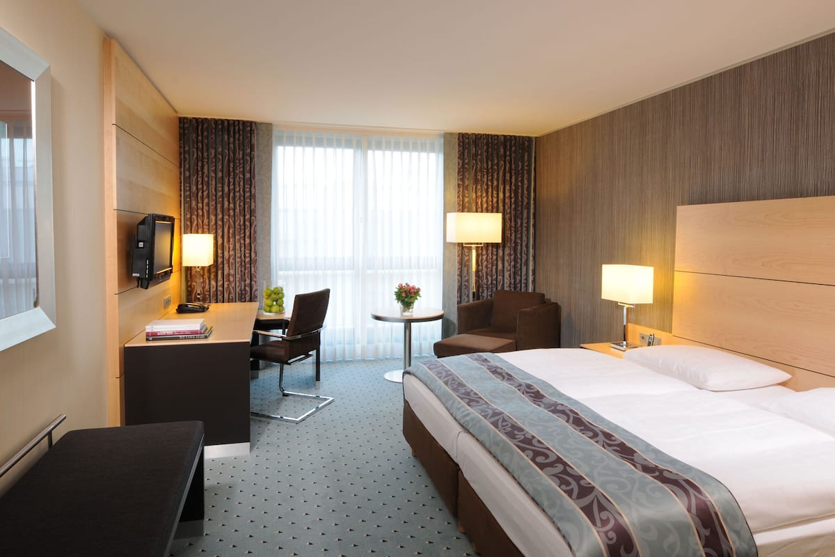 A hotel room with a large bed, a desk with a computer, a chair, a small table with a lamp, and a TV on the wall.