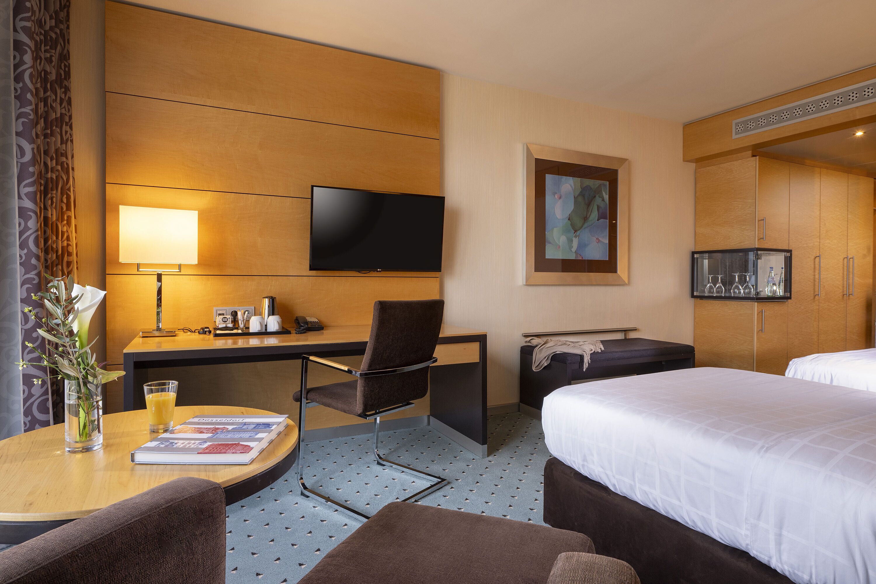 Ein Hotelzimmer mit Schreibtisch, Stuhl, Flachbildfernseher, Bett und einem kleinen Tisch mit Zeitschrift und Glas Orangensaft.