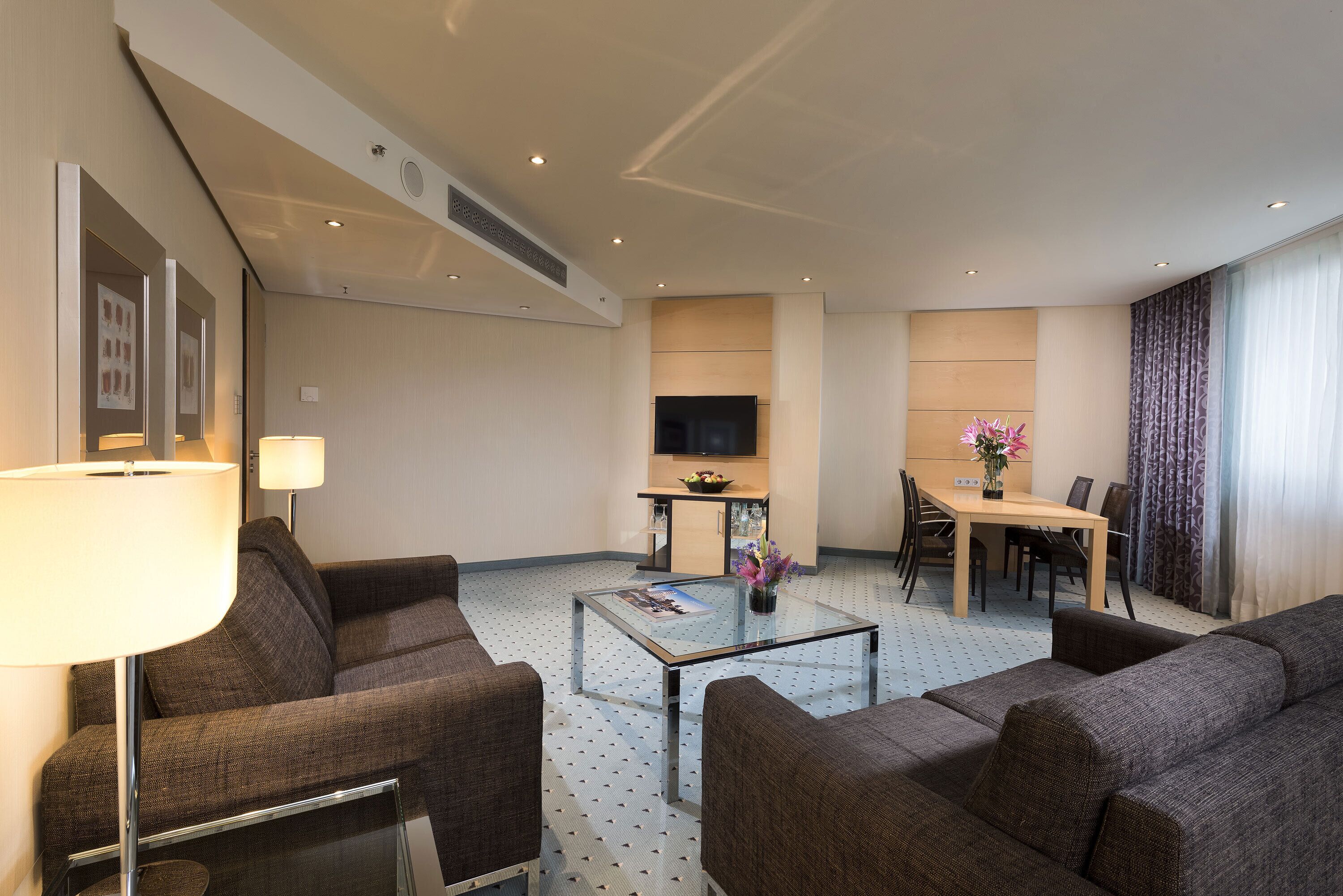 Une chambre d’hôtel équipée d’un canapé, d’une table basse, d’un téléviseur, d’une table à manger avec des chaises et d’un vase à fleurs.