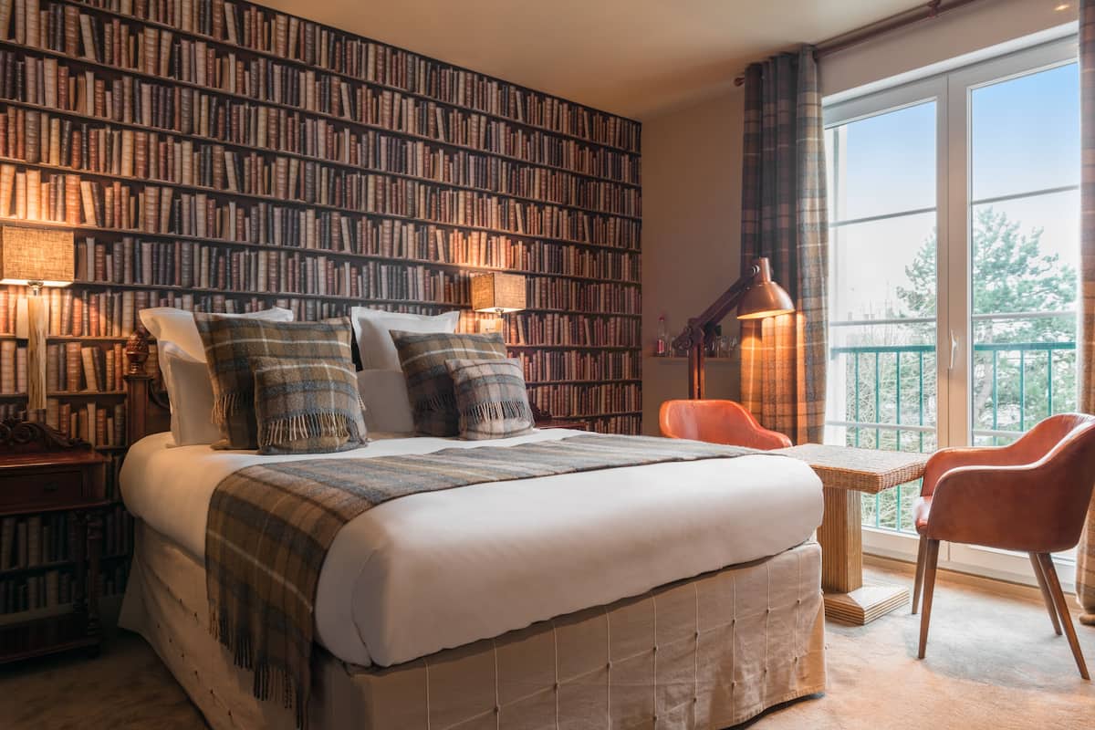 A bedroom with a large bookshelf, a bed with a white comforter, and a view of the outdoors through a window.