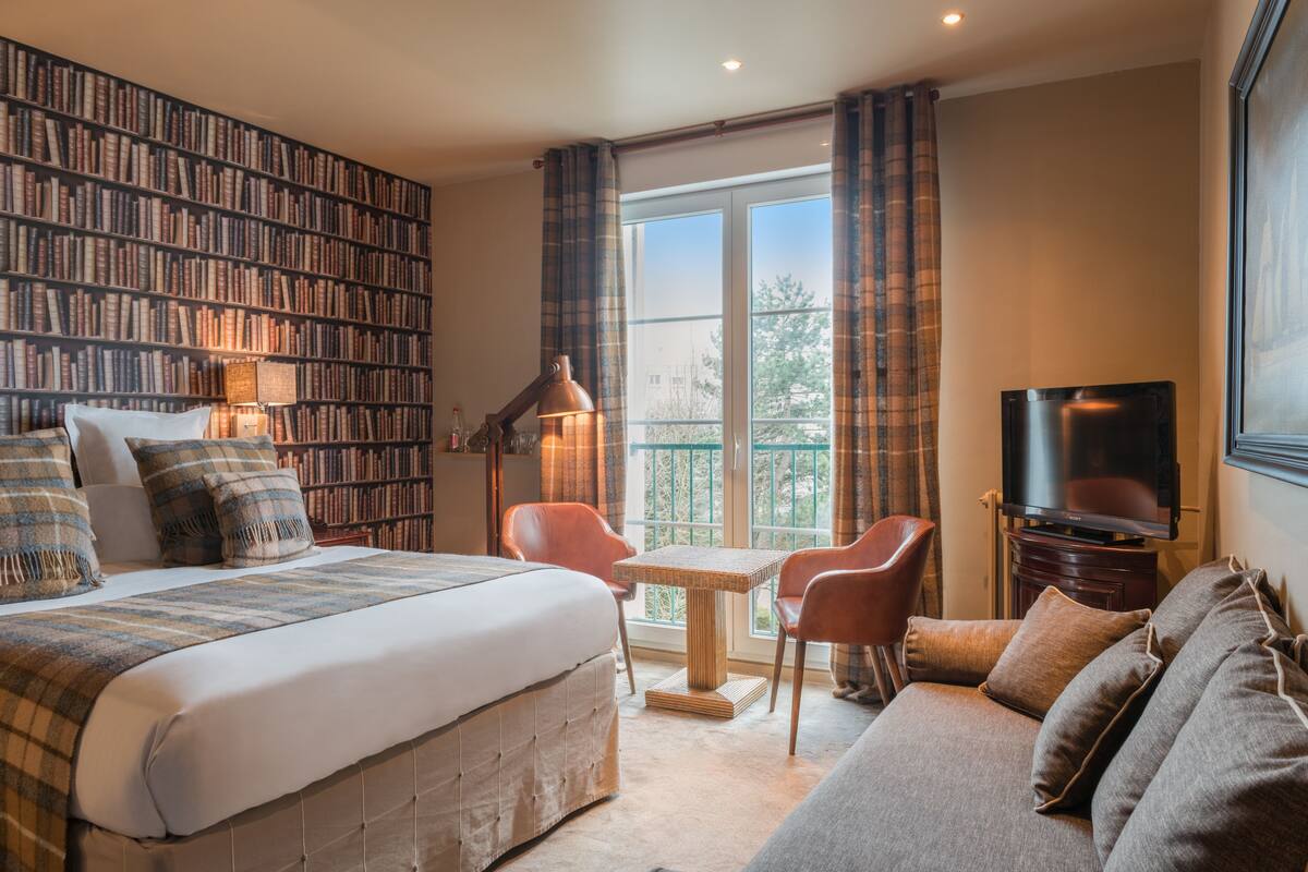 A hotel room with a large bookshelf, a bed, a sofa, a TV, and a small table.