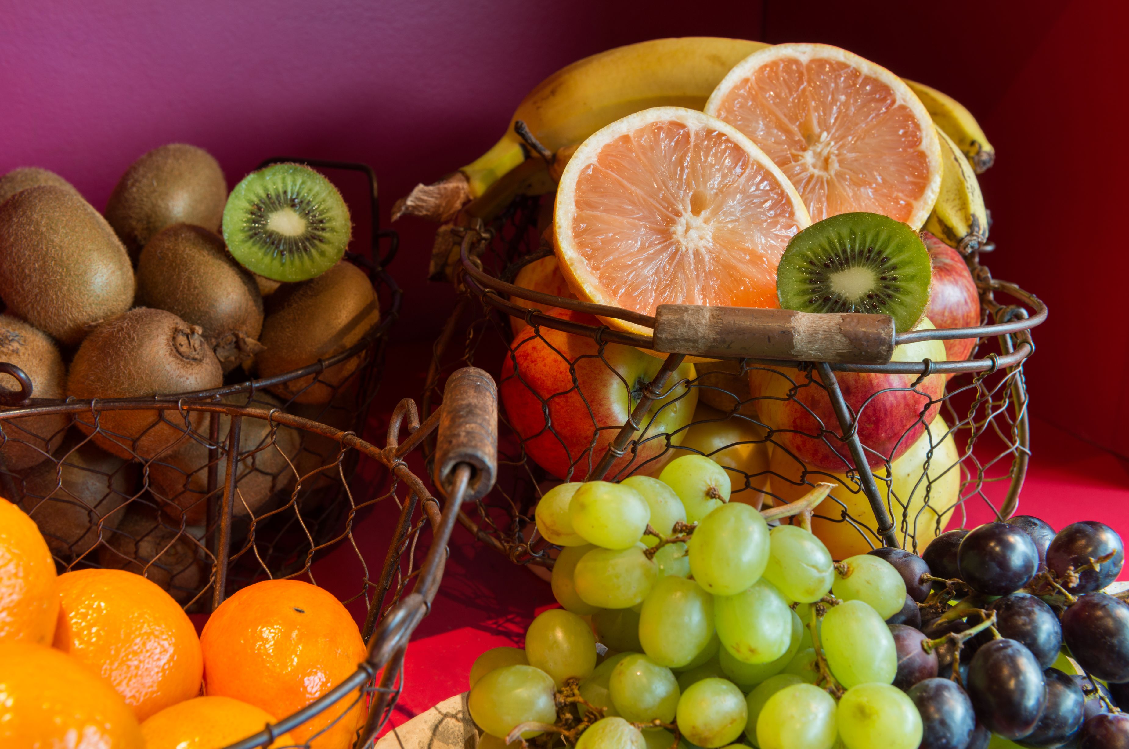 Dos cestas de alambre llenas de diversas frutas, como kiwis, naranjas, uvas y plátanos.
