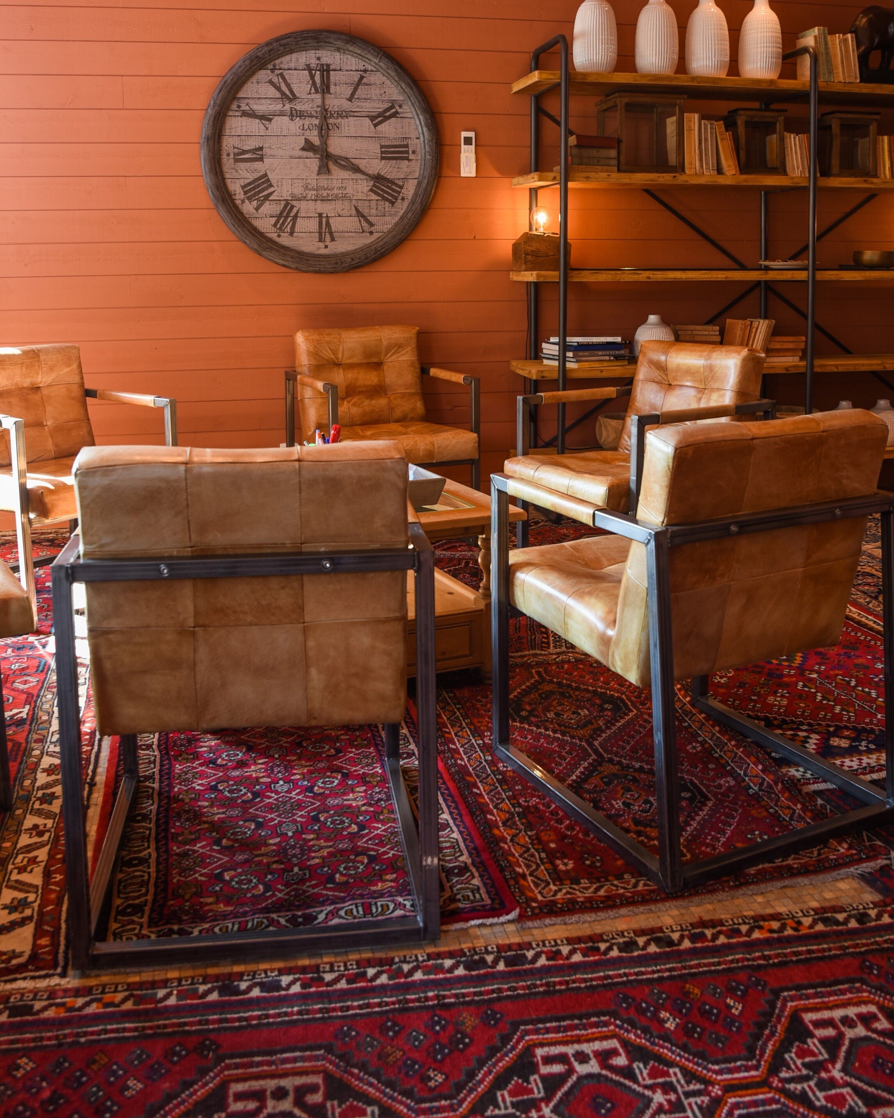 Een kamer met een grote wandklok, leren fauteuils, een kamerbreed tapijt en een boekenkast met boeken en decoratieve objecten.