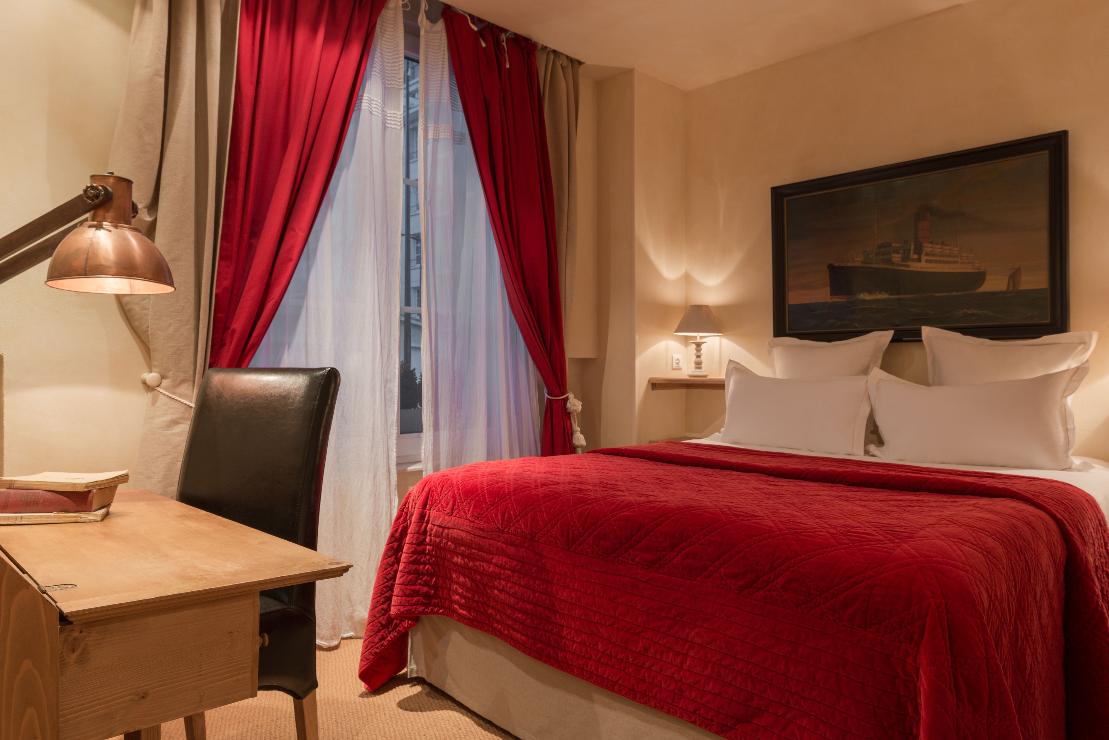 A hotel room with a bed, a desk, a chair, a lamp, and a painting on the wall.