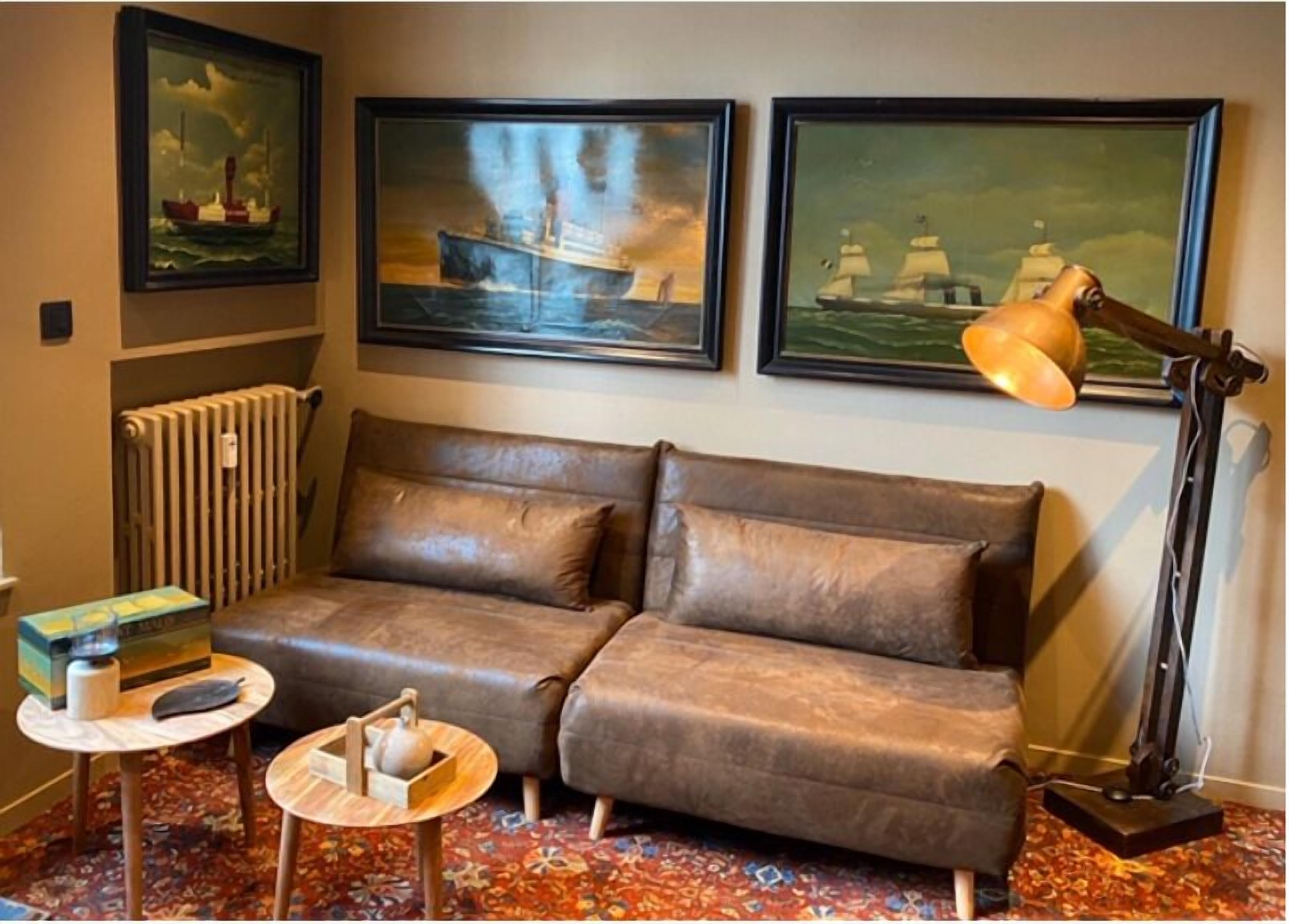 Una habitación con un sofá de cuero marrón, una mesita redonda con una tetera, un radiador y tres cuadros enmarcados de barcos en la pared.