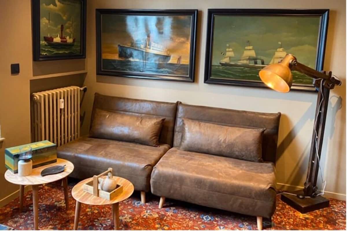 A room with a brown leather sofa, a small round table with a teapot, a radiator, and three framed paintings of ships on the wall.