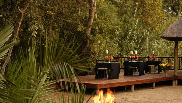 Un área de comedor al aire libre con un brasero, mesas y sillas sobre una terraza de madera rodeada de exuberante vegetación.