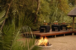 Un espace de restauration en plein air avec un foyer, des tables et des chaises disposées sur une terrasse en bois, le tout entouré d’une végétation luxuriante.