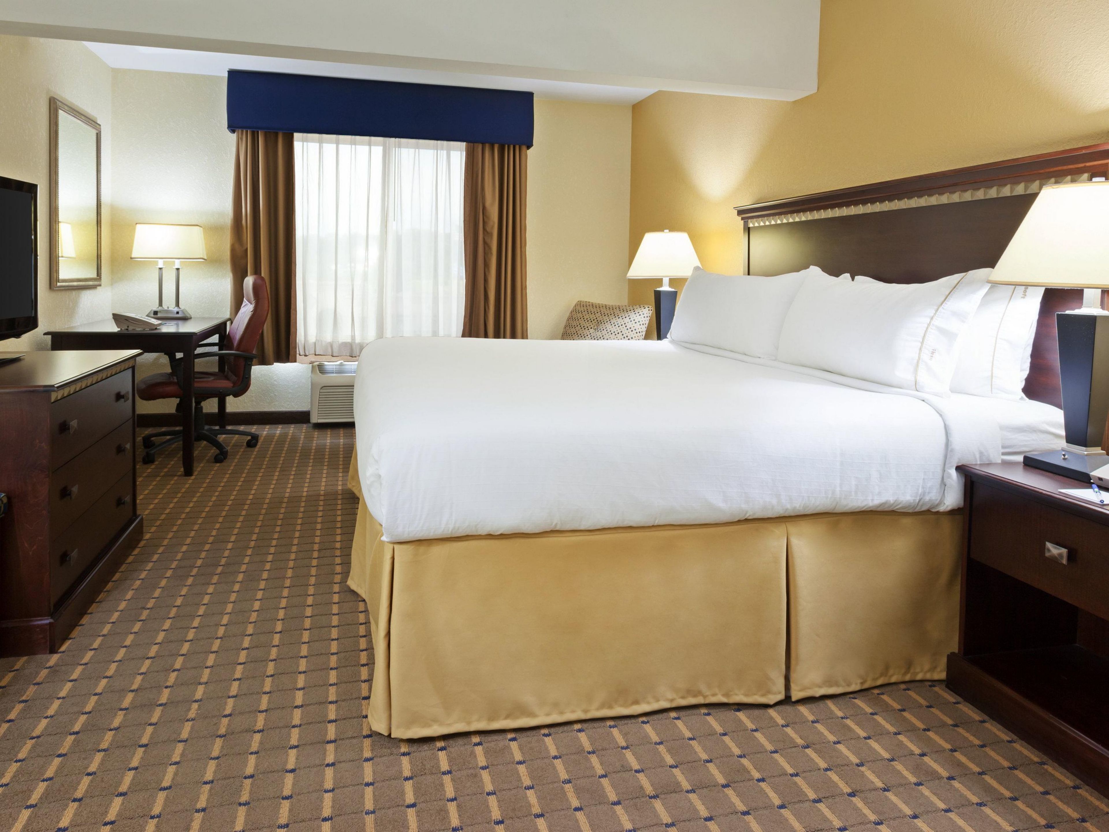 Ein Hotelzimmer mit einem großen Bett, einem Schreibtisch mit Stuhl, einem Fernseher und einem Fenster mit Vorhängen.