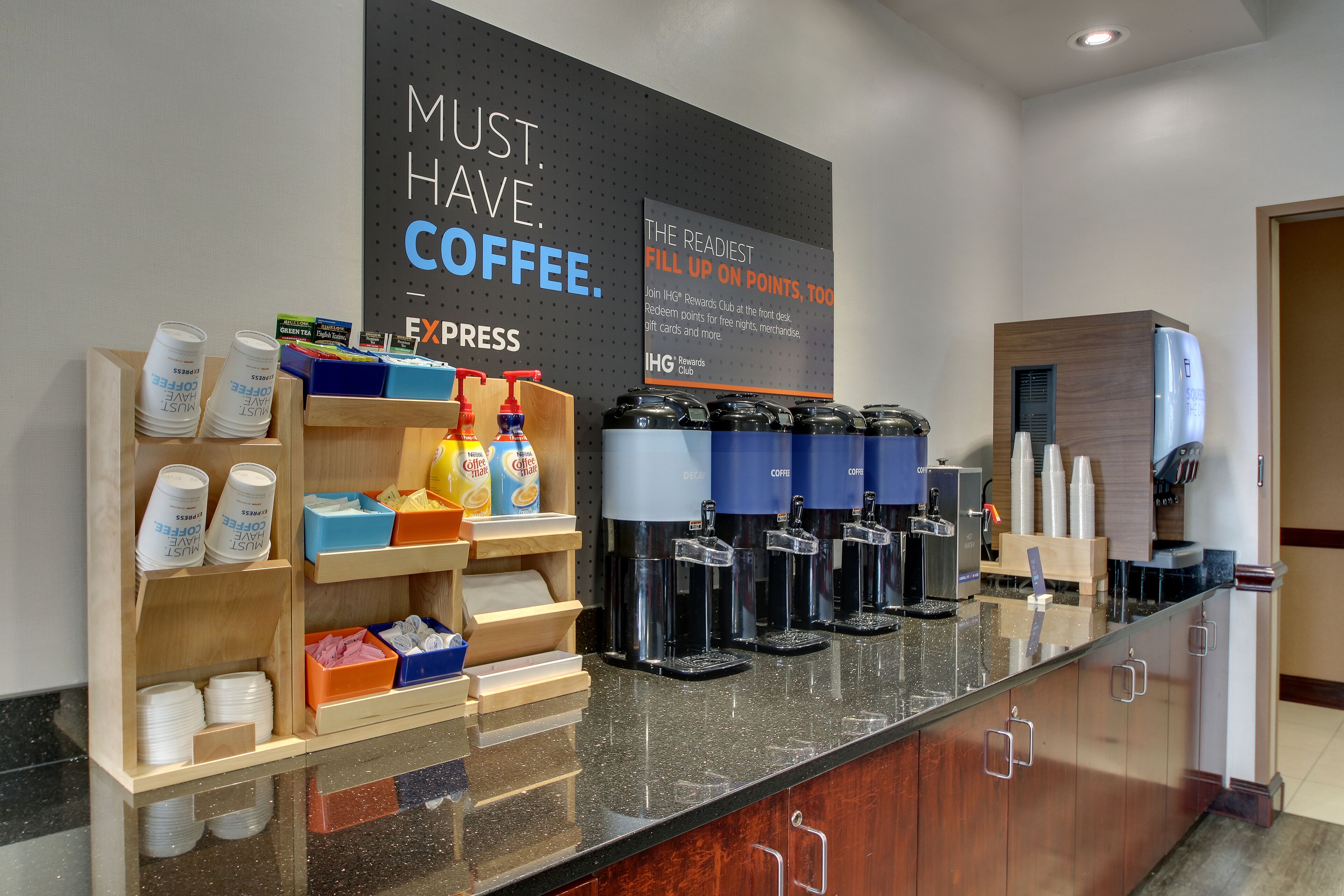 Een koffiehoek met verschillende koffieapparaten, een bord dat de IHG Rewards Club promoot, en diverse koffiekopjes en -bekers.