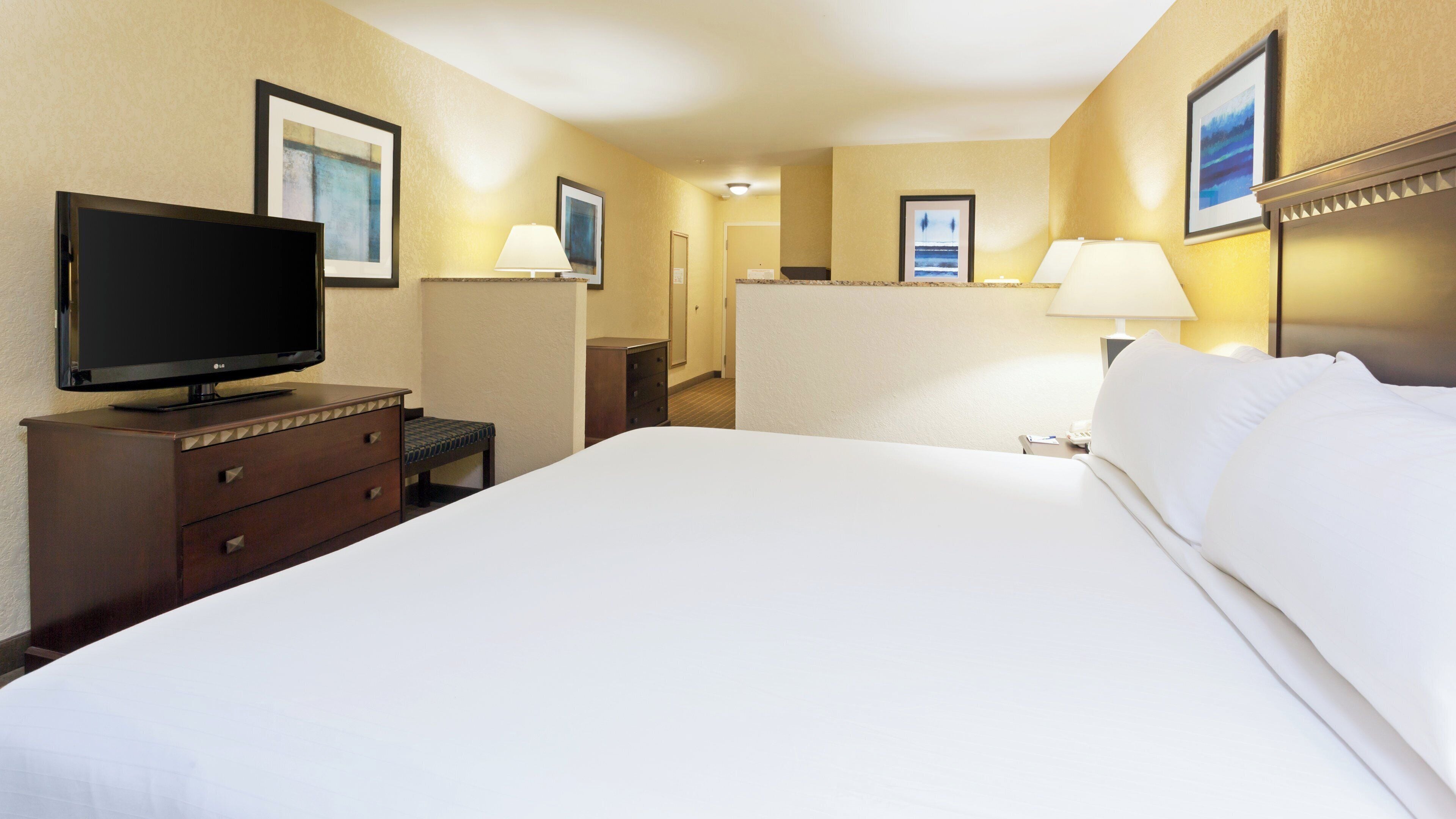 A hotel room with a bed, a television, a desk with drawers, a lamp, and framed pictures on the walls.