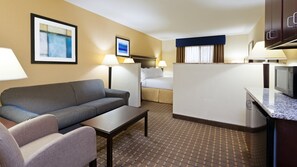 A hotel room with a sofa, armchair, a small table, a bed, a desk with a computer, and a microwave.
