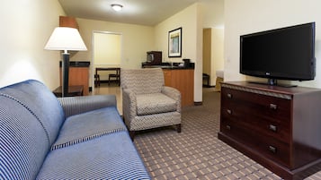 Habitación de hotel con un sofá a rayas, un sillón estampado, un televisor sobre un mueble de madera y una kitchenette al fondo.
