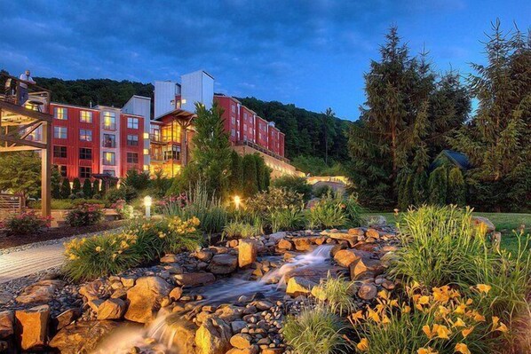 A hotel with multiple red buildings, a landscaped garden with a small stream, and a forested hillside in the background.