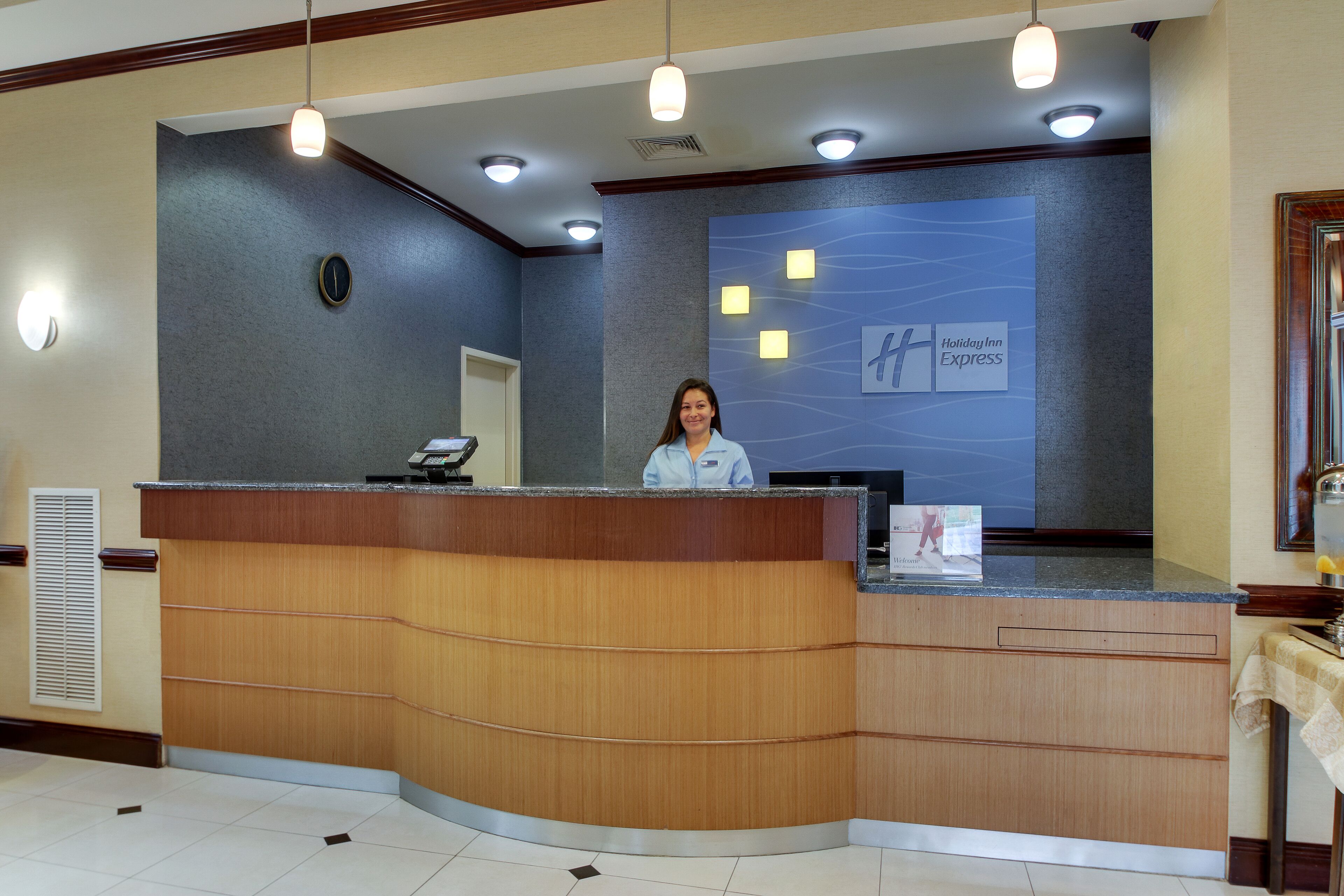 Un mostrador de recepción con una mujer detrás, un letrero de Holiday Inn Express en la pared y una pequeña mesa con mantel a la derecha.