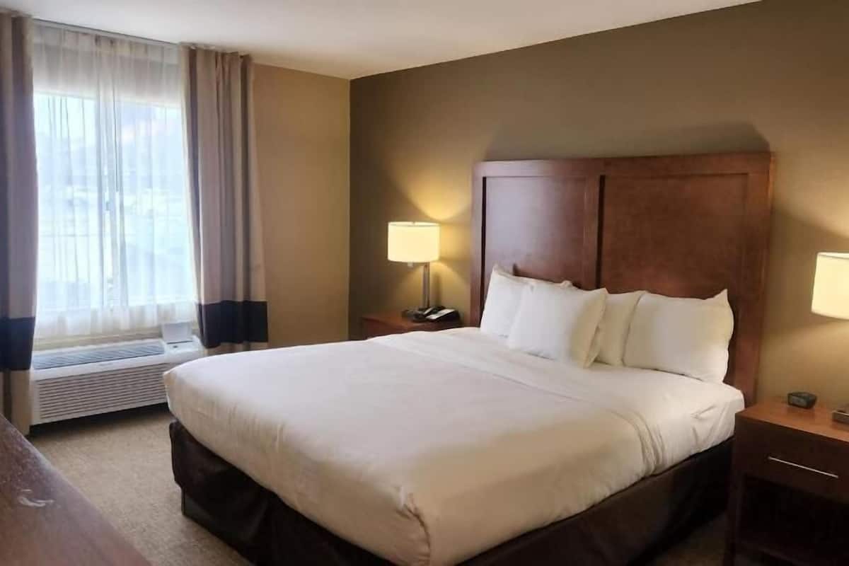 Una habitación de hotel con una cama grande, dos mesitas de noche con lámparas, una ventana con cortinas y un calentador fijado a la pared.