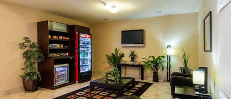 A hotel lobby with a vending machine, seating area, potted plants, and a flat-screen TV.