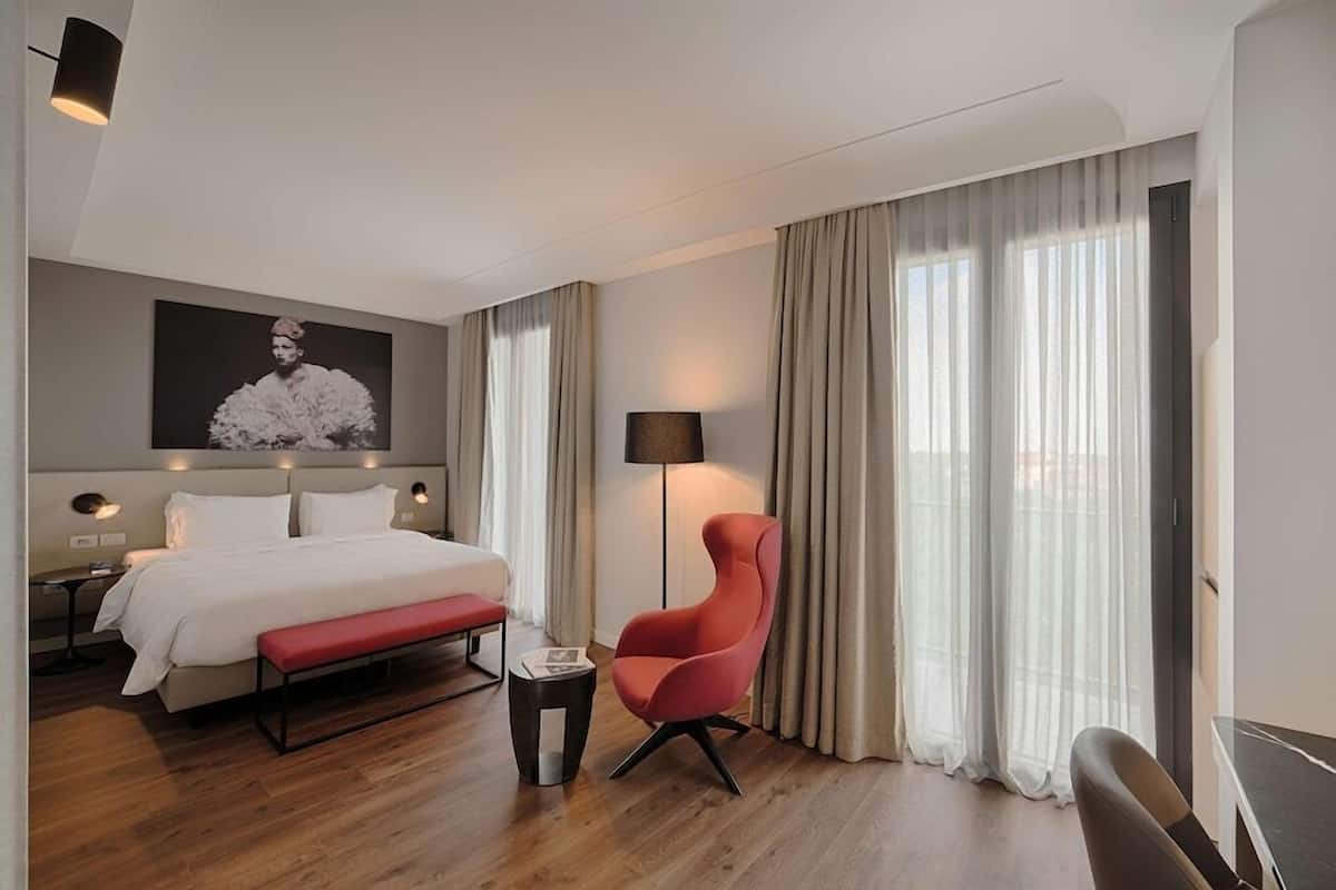 A hotel room with a bed, a red armchair, a bedside table, a lamp, and a large window with curtains.