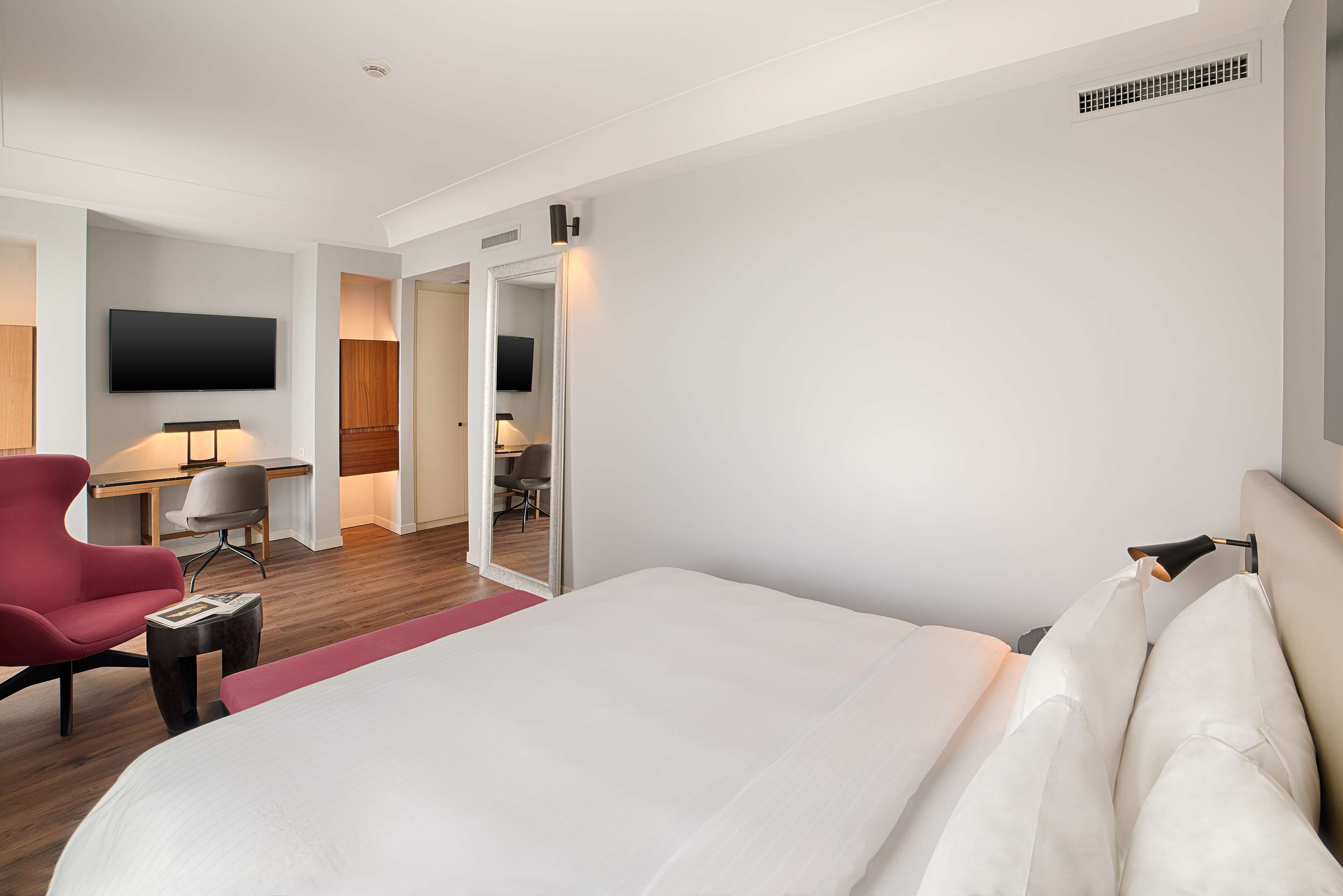 Camera d'albergo con un letto grande, una poltrona rossa, una scrivania con una sedia, una TV fissata al muro e una porta che dà su un'altra stanza.